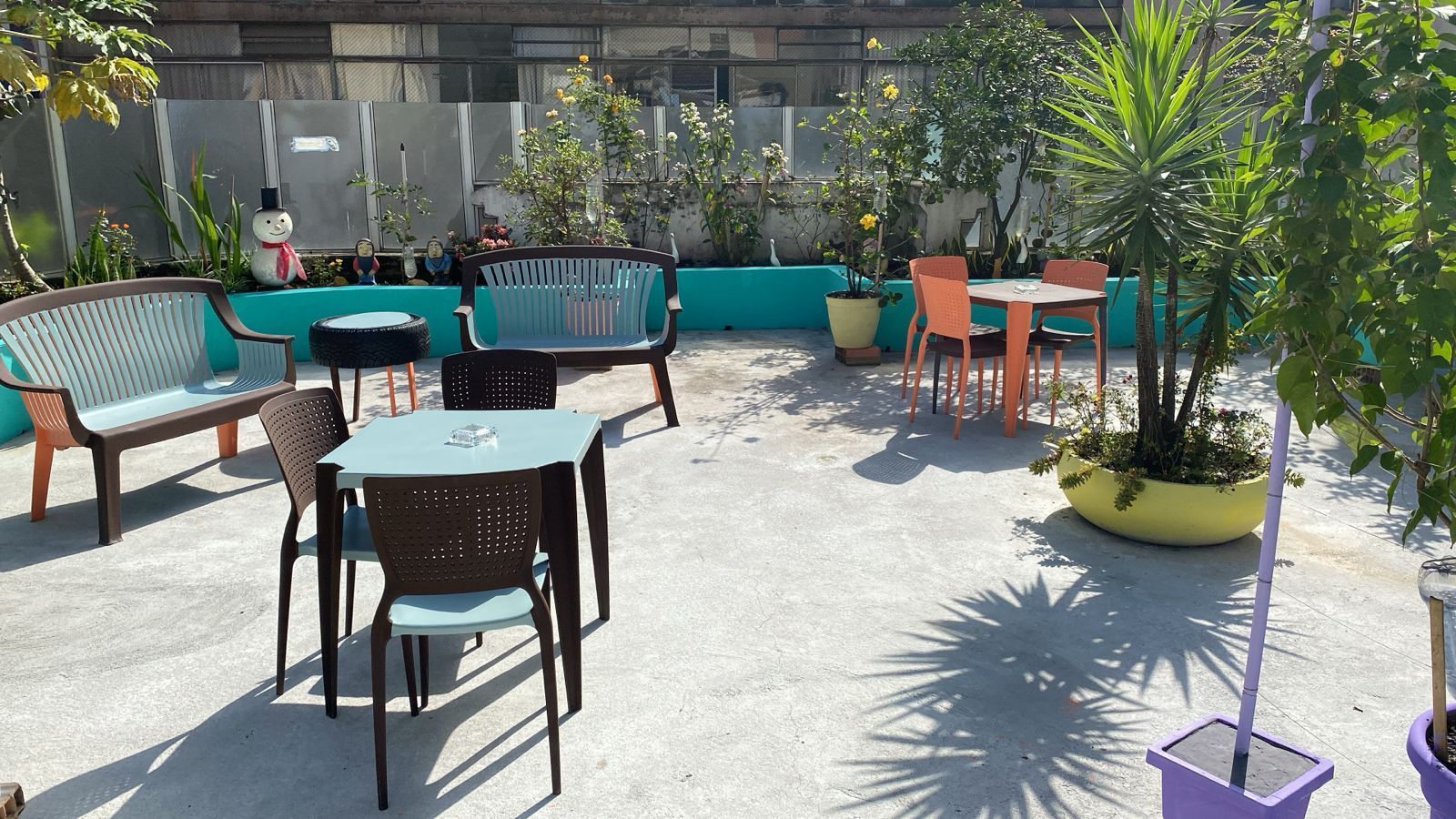 Mesas de café ao ar livre com bancos turquesa, mesas pretas e vasos de plantas sob a luz do sol.