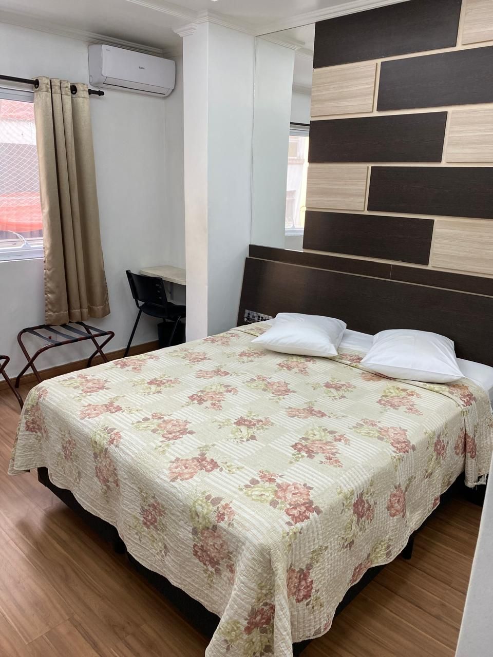 Quarto pequeno com cama de casal, colcha floral, travesseiros, ar-condicionado de parede e uma cadeira junto à janela.