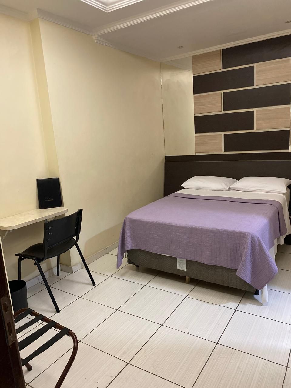Quarto pequeno com cama, escrivaninha, cadeira preta e piso de azulejo em um interior moderno e minimalista.
