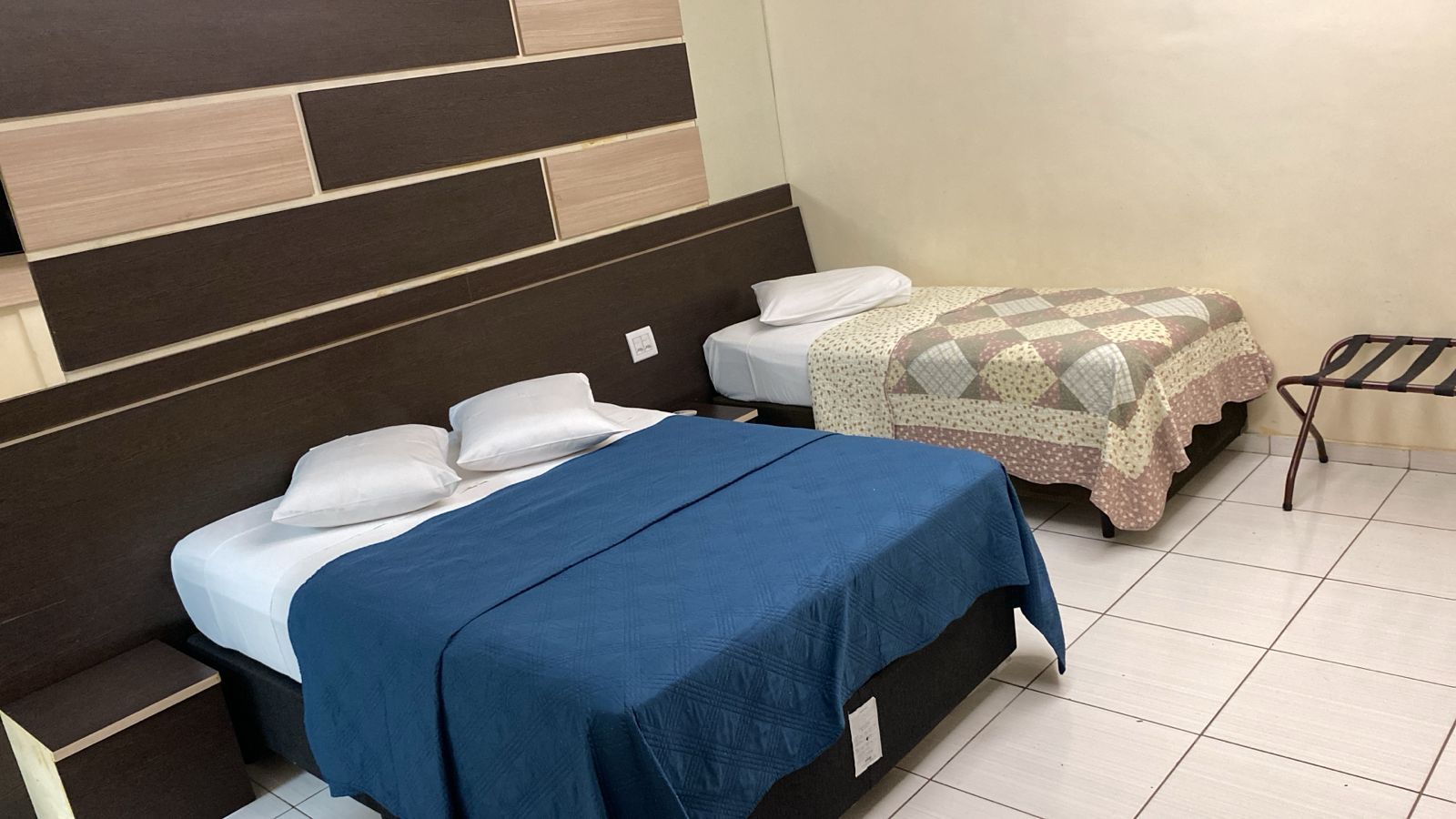 Quarto pequeno com duas camas, uma com lençóis azuis e outra estampada, sobre piso de azulejos brancos.
