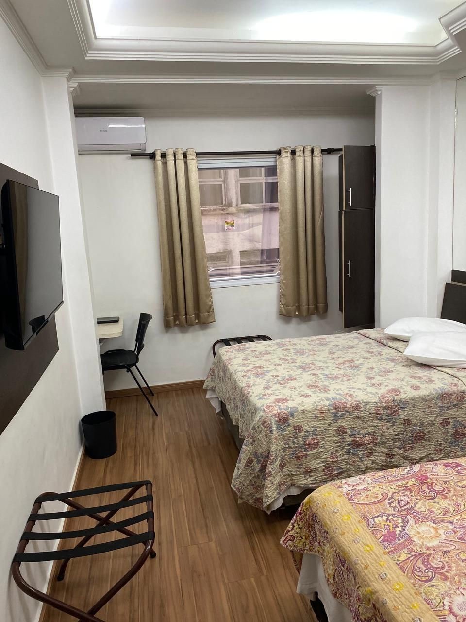 Quarto de hotel pequeno com duas camas, cortinas nas janelas, TV de parede e piso de madeira.