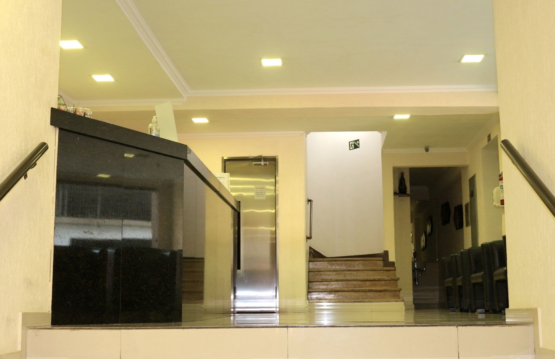 Hall de entrada com escadas, elevador espelhado e corrimãos pretos sob luzes de teto em tons quentes.