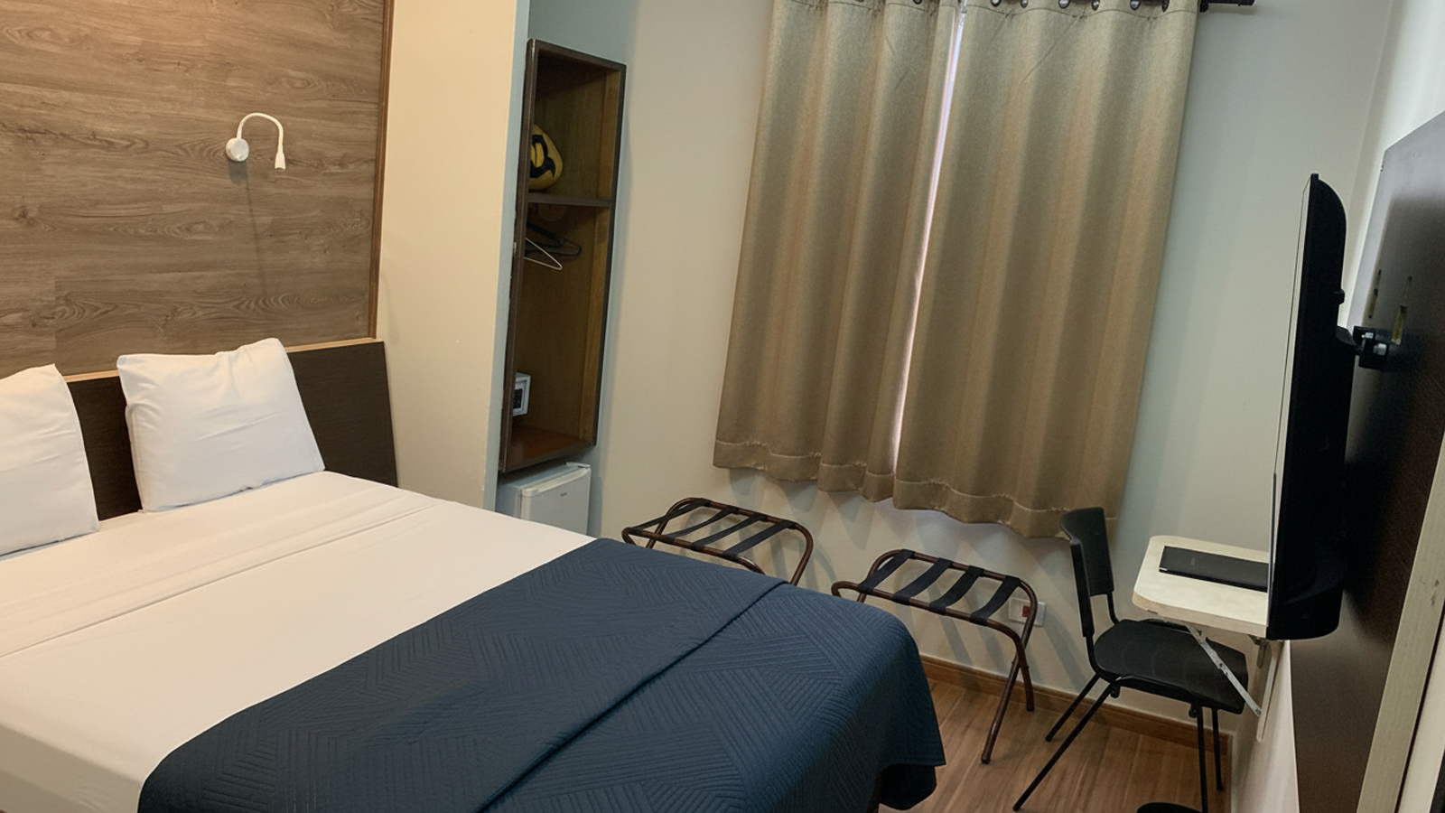 Quarto de hotel pequeno com cama, cortinas, TV, cadeira e suporte para bagagem.
