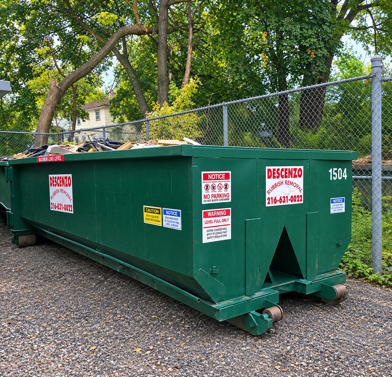 Large green roll-off dumpster positioned on gravel beside a chain-link fence.