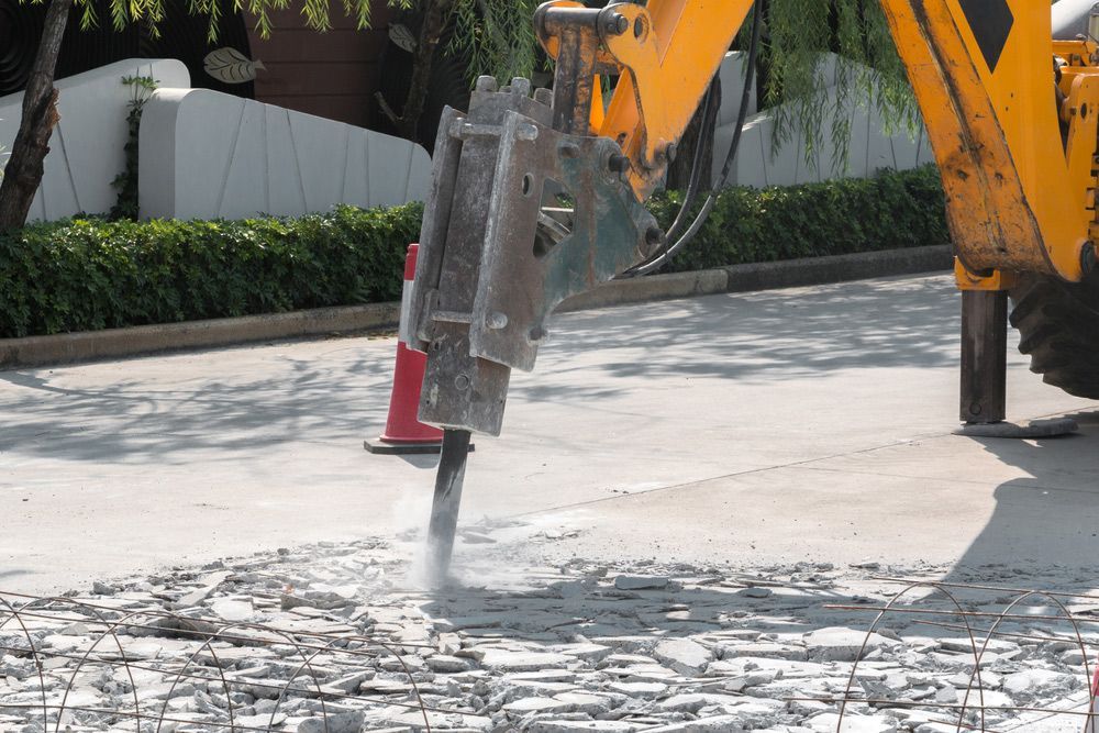 A yellow excavator is breaking concrete on a street