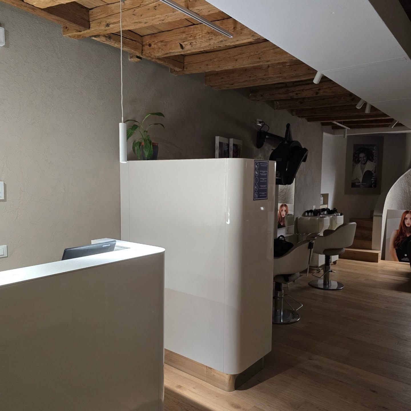 Interno di un salone moderno con postazioni di lavoro bianche, soffitto con travi a vista e una cliente seduta sullo sfondo.