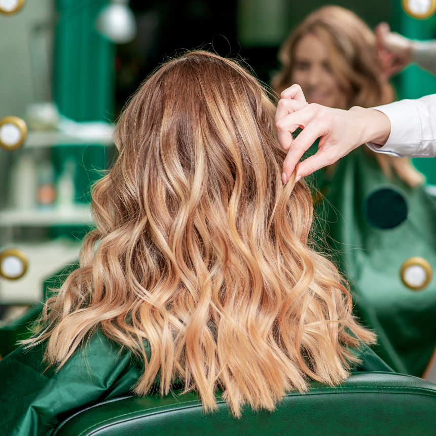 Capelli biondi ondulati acconciati su una poltrona da salone verde, con un parrucchiere che sistema le ciocche davanti a uno specchio.