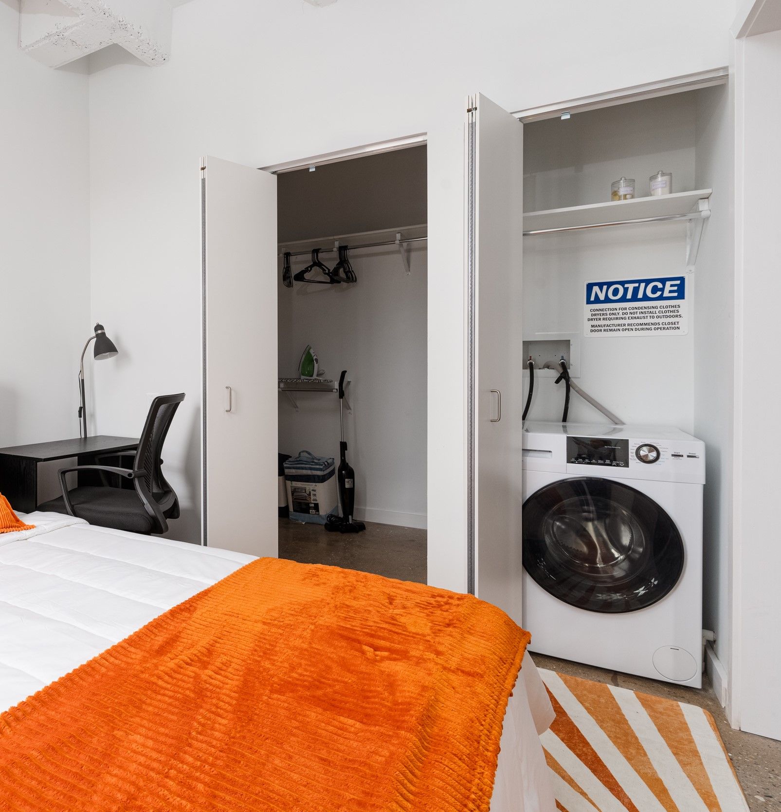 A bedroom with a washing machine and closet.  White walls and closet doors frame the washer. An orange blanket adorns the bed.