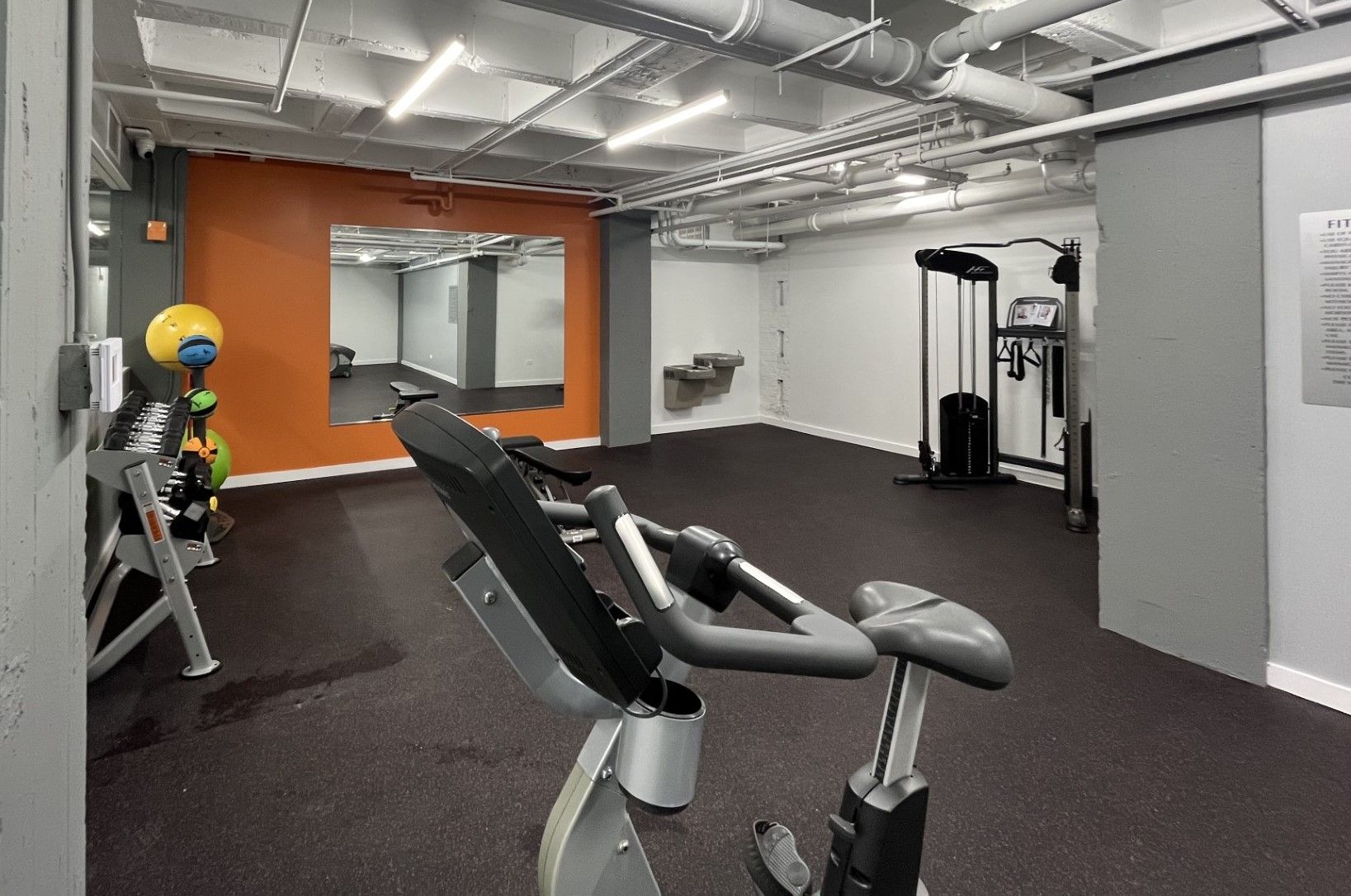 Gym interior with exercise bike in foreground, weights and equipment in the background. Orange and gray walls, black floor, and exposed ceiling.