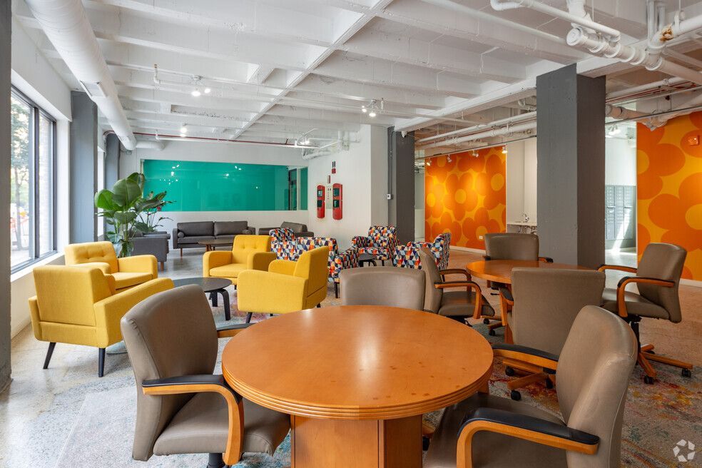 A colorful co-working space with round tables, armchairs, and couches. The walls are orange and turquoise, and there are large windows.