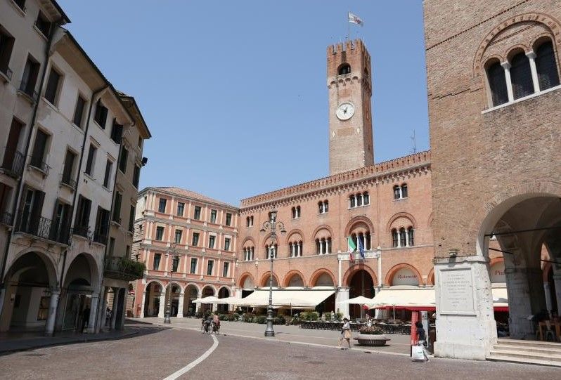 Piazza storica con torre dell'orologio in mattoni, portici e ombrelloni da caffè sotto un cielo azzurro e limpido.
