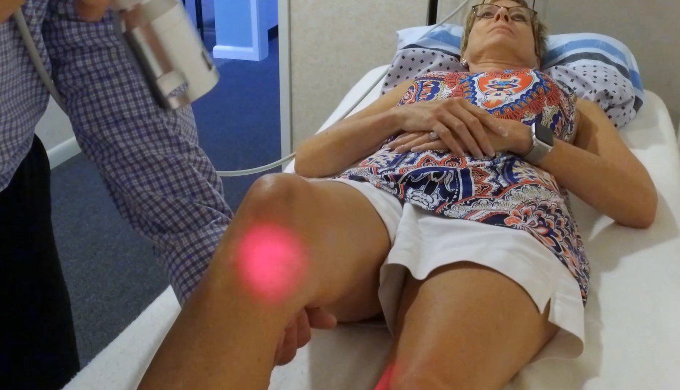 A person receives laser therapy on their knee while lying on an examination table. A red light is visible in Hope Mills, NC.