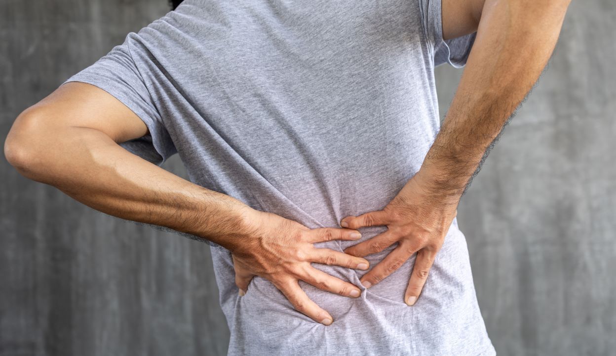 Man clutching lower back in pain, grey shirt, against grey wall in Hope Mills, NC.
