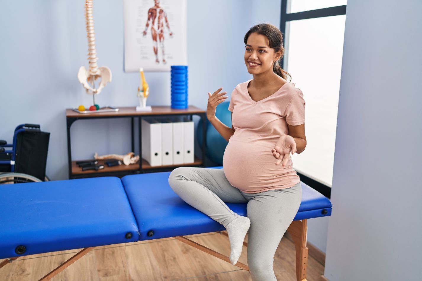 Pregnant woman sitting on a blue examination table in a medical office, smiling and gesturing in Hope Mills, NC.