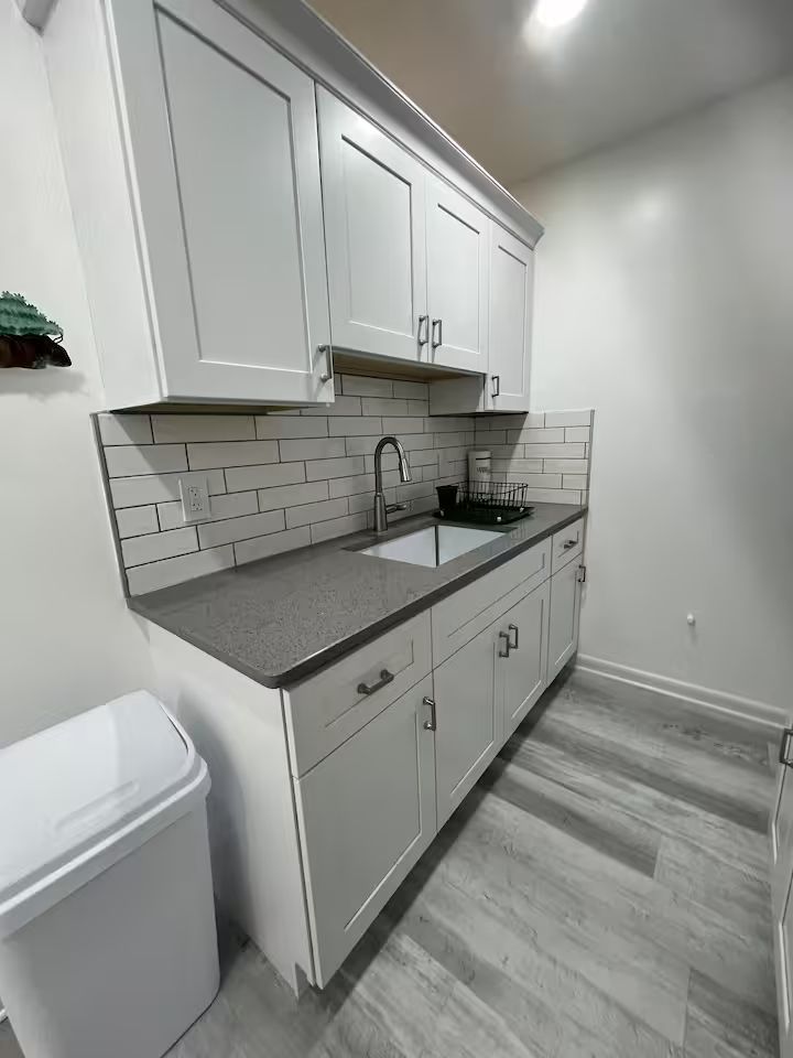 A kitchen with white cabinets , a sink , and a trash can.