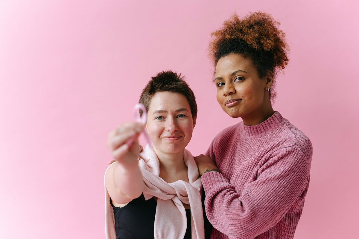Two women on pink background, one holding a pink ribbon, supporting breast cancer awareness.