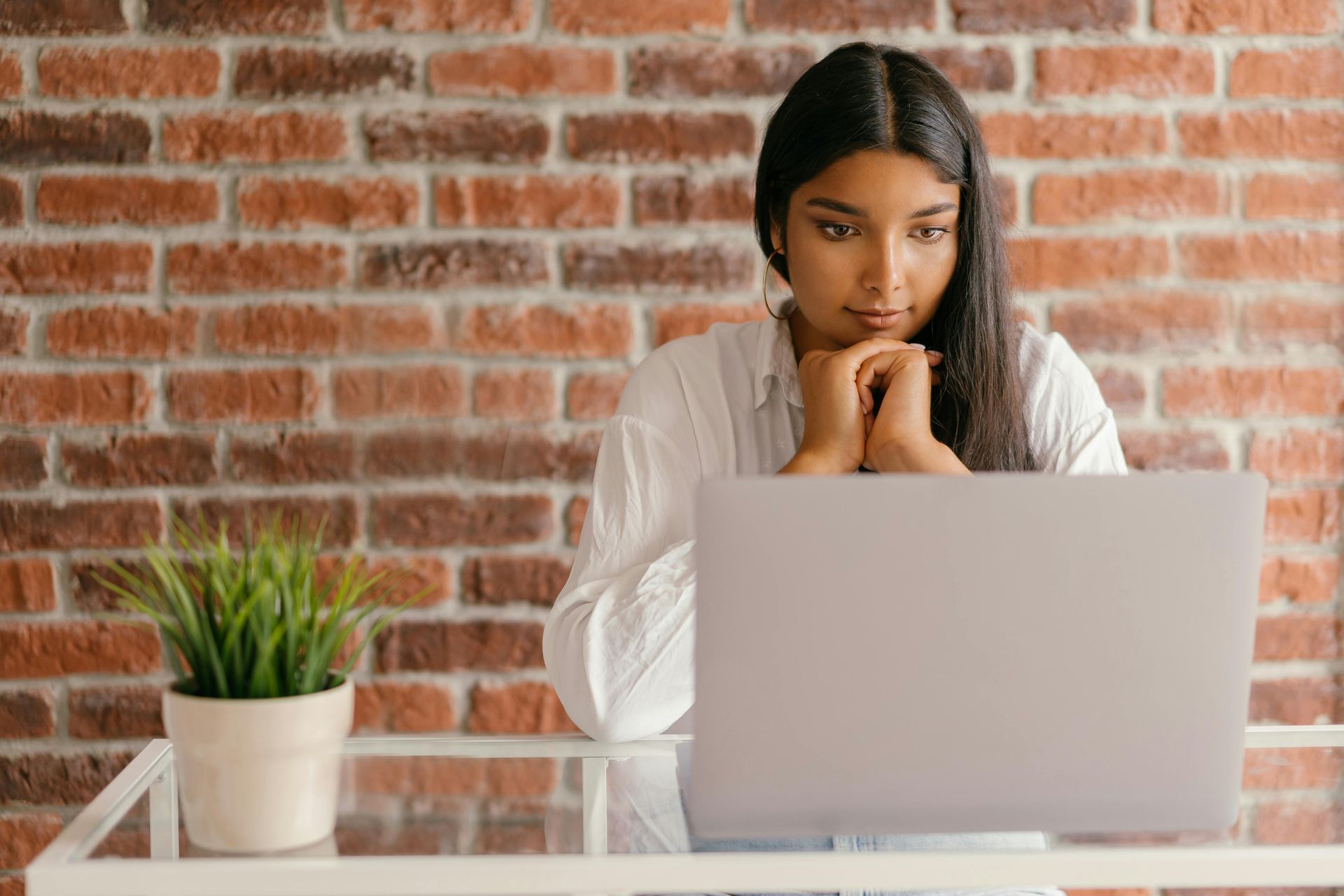 Woman in white shirt, hands clasped, focused on laptop. Brick wall background, potted plant.