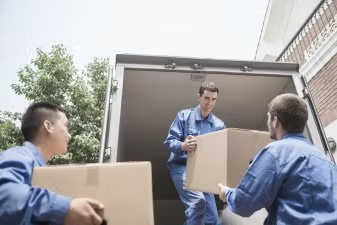 Movers in blue uniforms unloading boxes from a truck outside a building