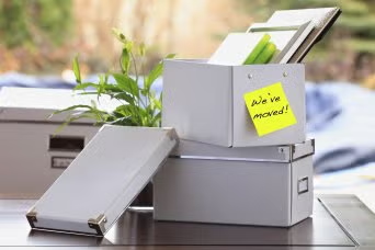 Moving boxes and a plant with a yellow “We’ve moved!” note on a desk