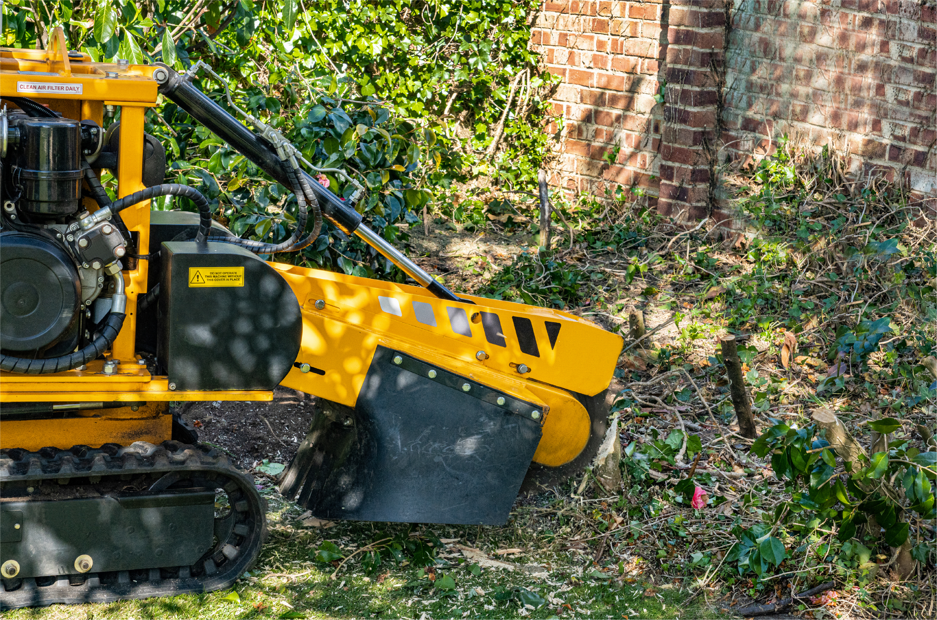 A yellow stump grinder is sitting in the grass next to a brick wall.