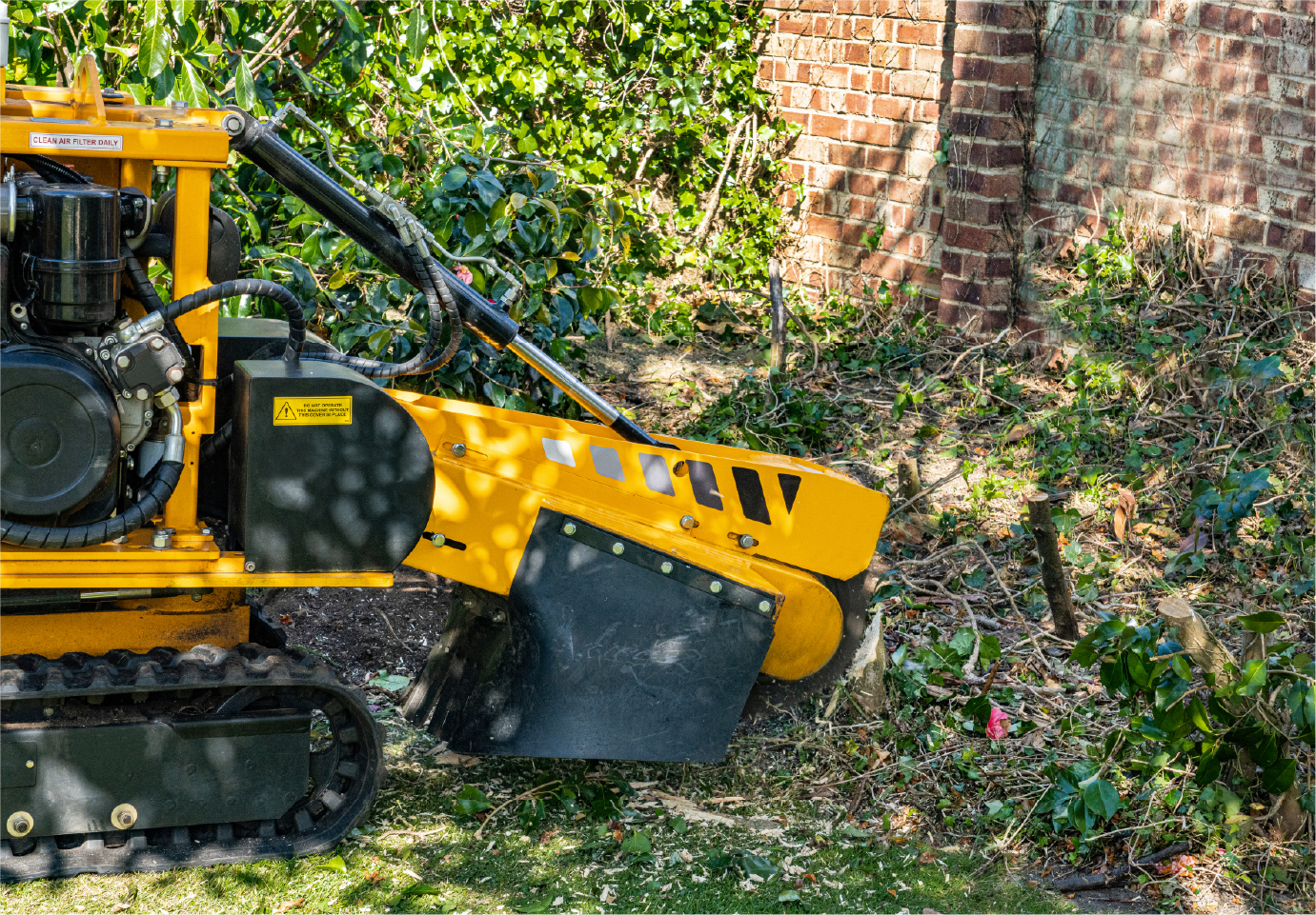 A yellow stump grinder is sitting in the grass next to a brick wall.