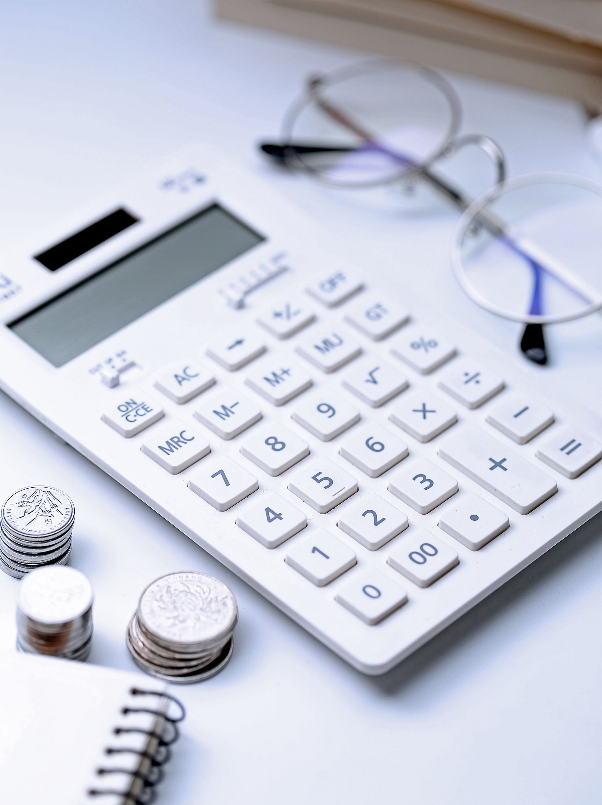 Une calculatrice blanche, des pièces de monnaie, des lunettes et un bloc-notes sur une surface blanche.