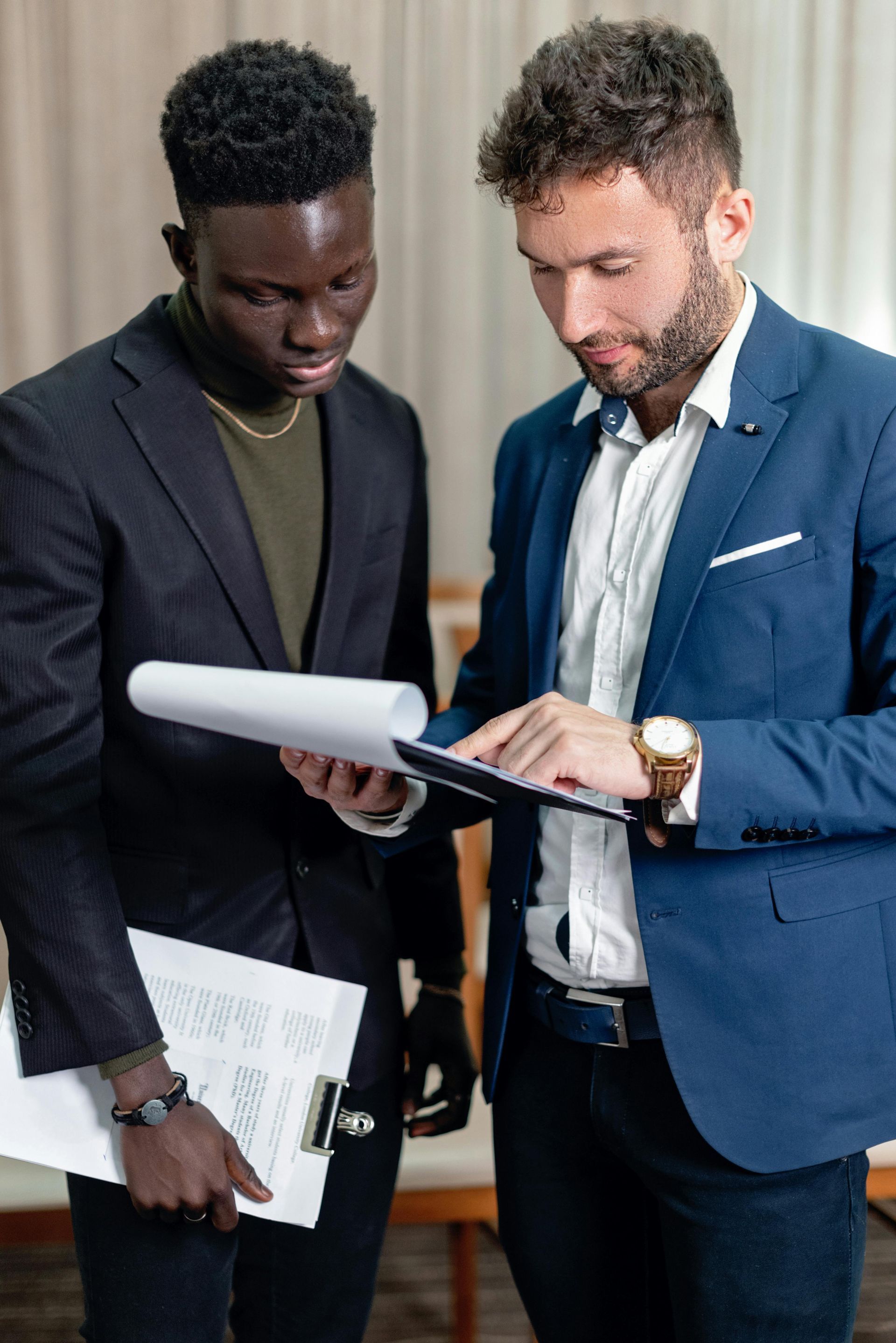 Two men in suits reviewing documents indoors, one points at a tablet.