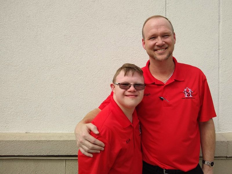 Two smiling people in red polos standing against a plain wall, one with an arm around the other.