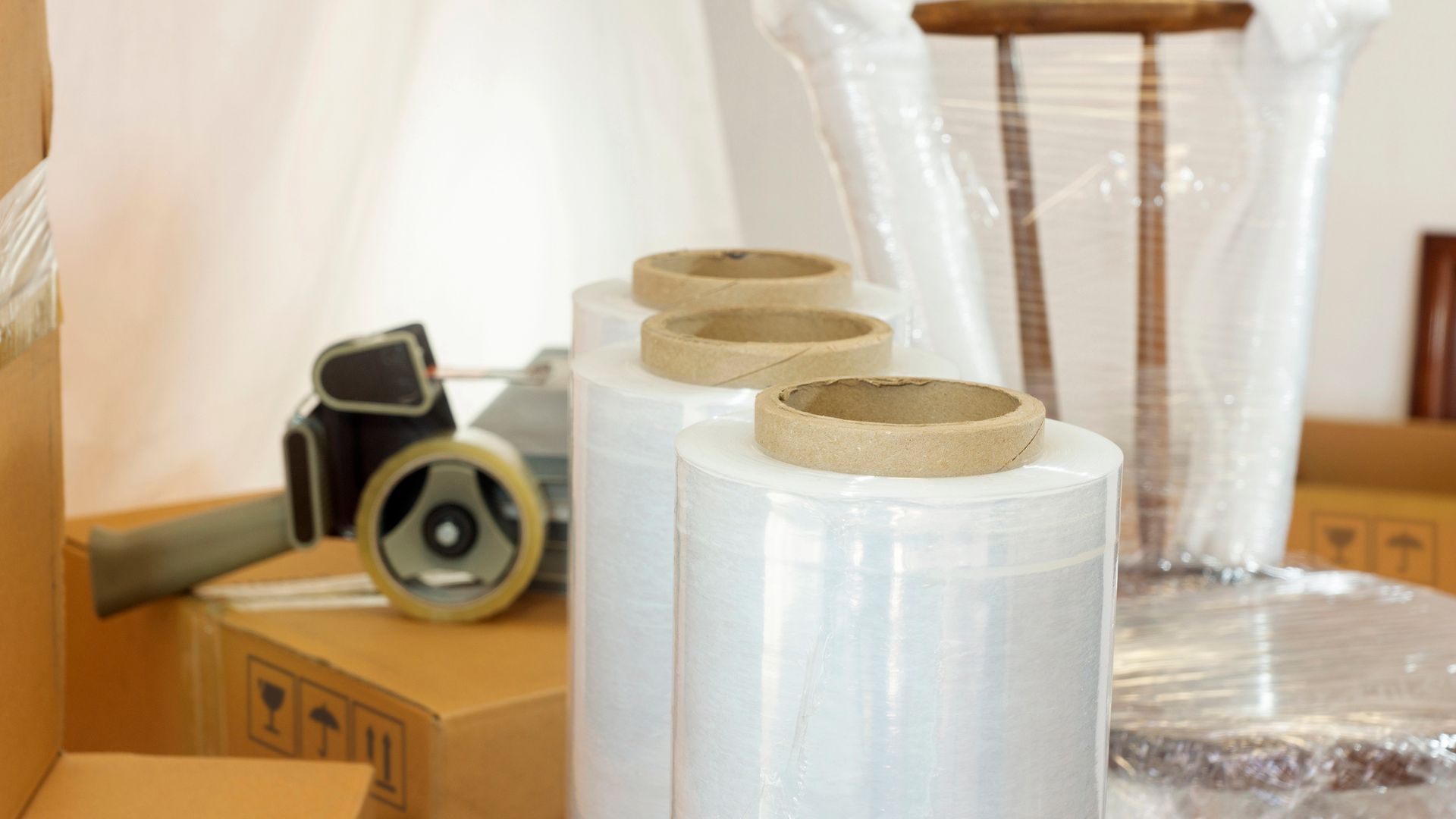 Materiale per il trasloco: rotoli di pellicola trasparente, scatole, dispenser per nastro adesivo e un tavolo rivestito.
