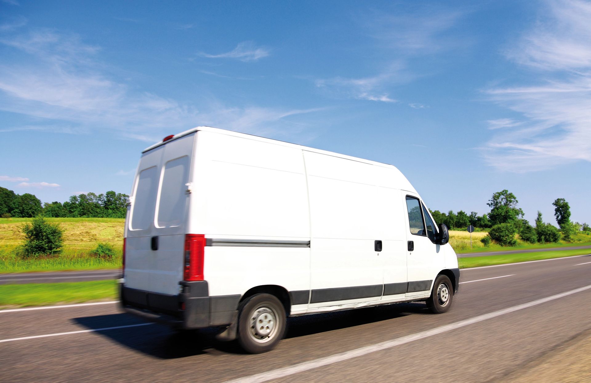 Un furgone bianco per merci viaggia in autostrada, sotto un cielo azzurro, attraversando campi verdi.