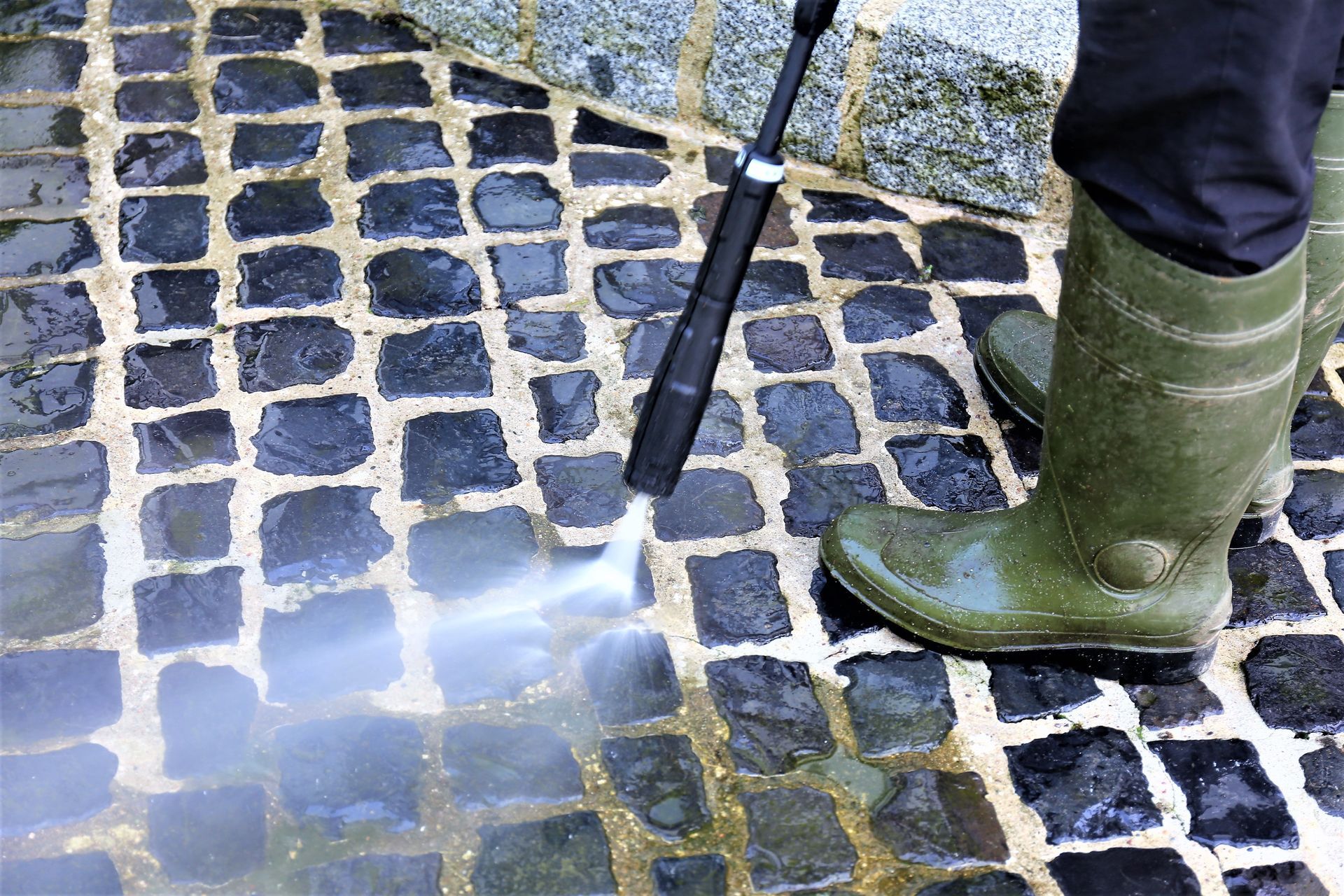 Person in green boots pressure washing a cobblestone walkway.