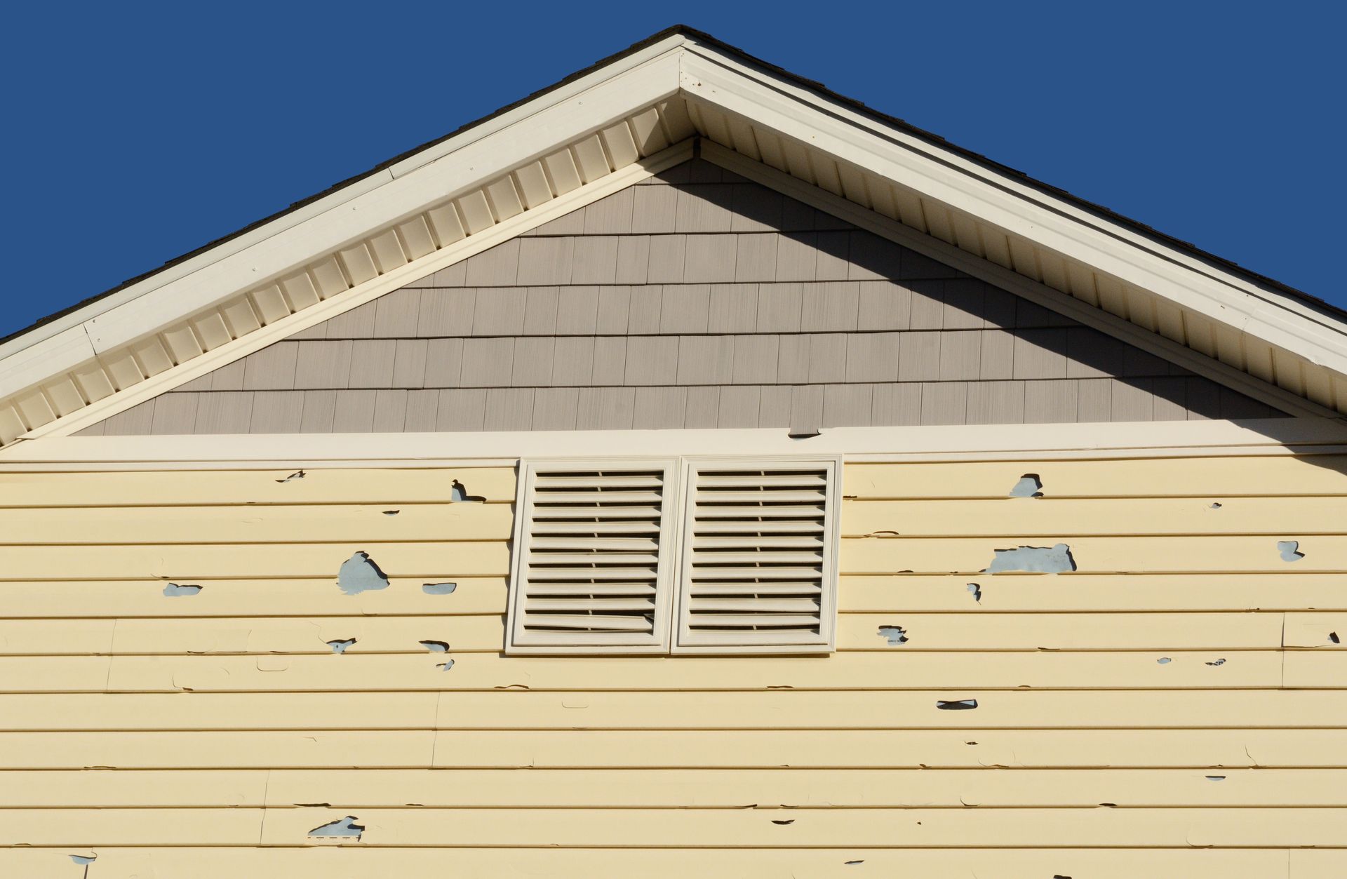 Yellow house siding with peeling paint, a vent, and a roof gable against a blue sky.