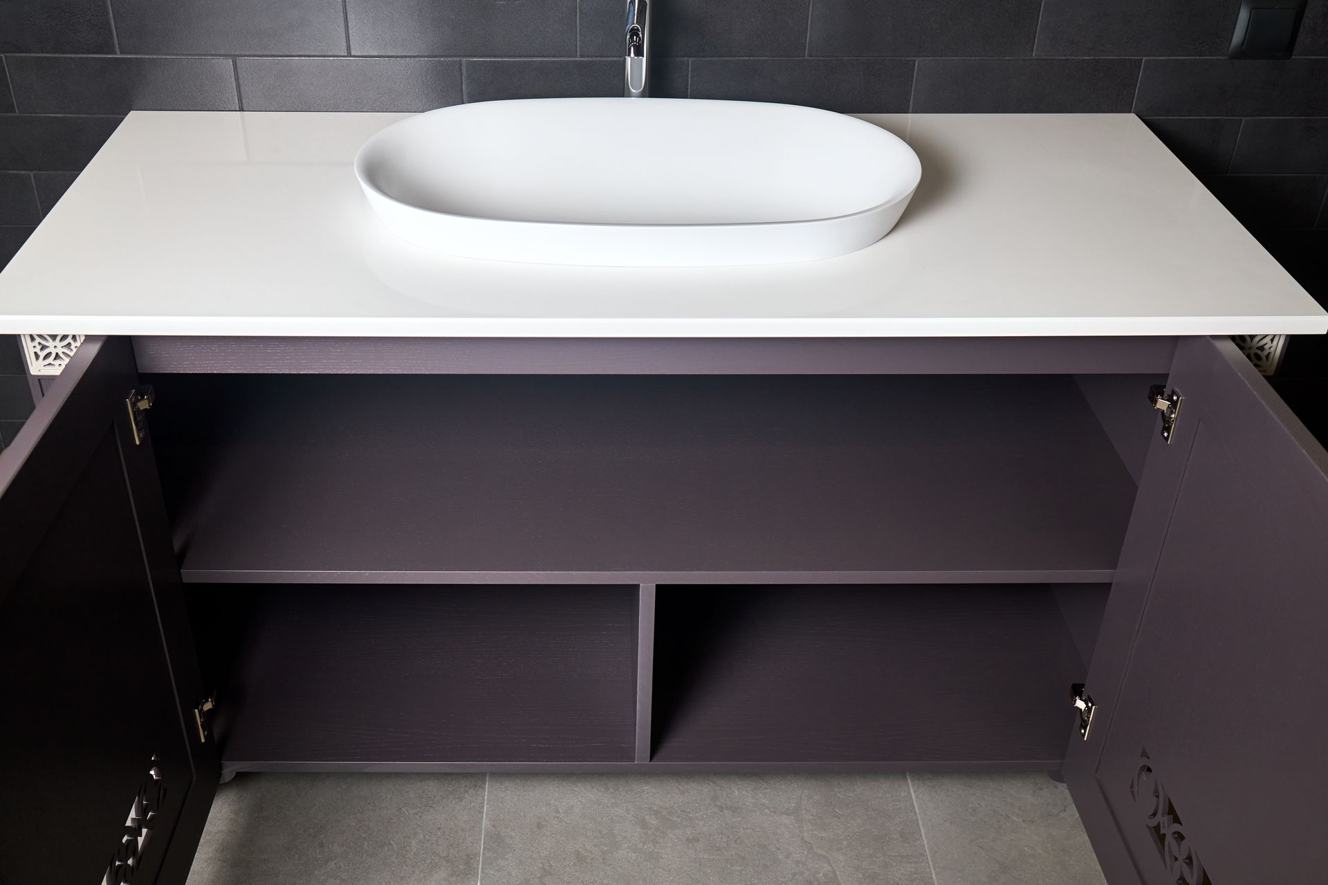 Bathroom vanity with a white countertop and oval sink, open to reveal shelves. Gray cabinets.