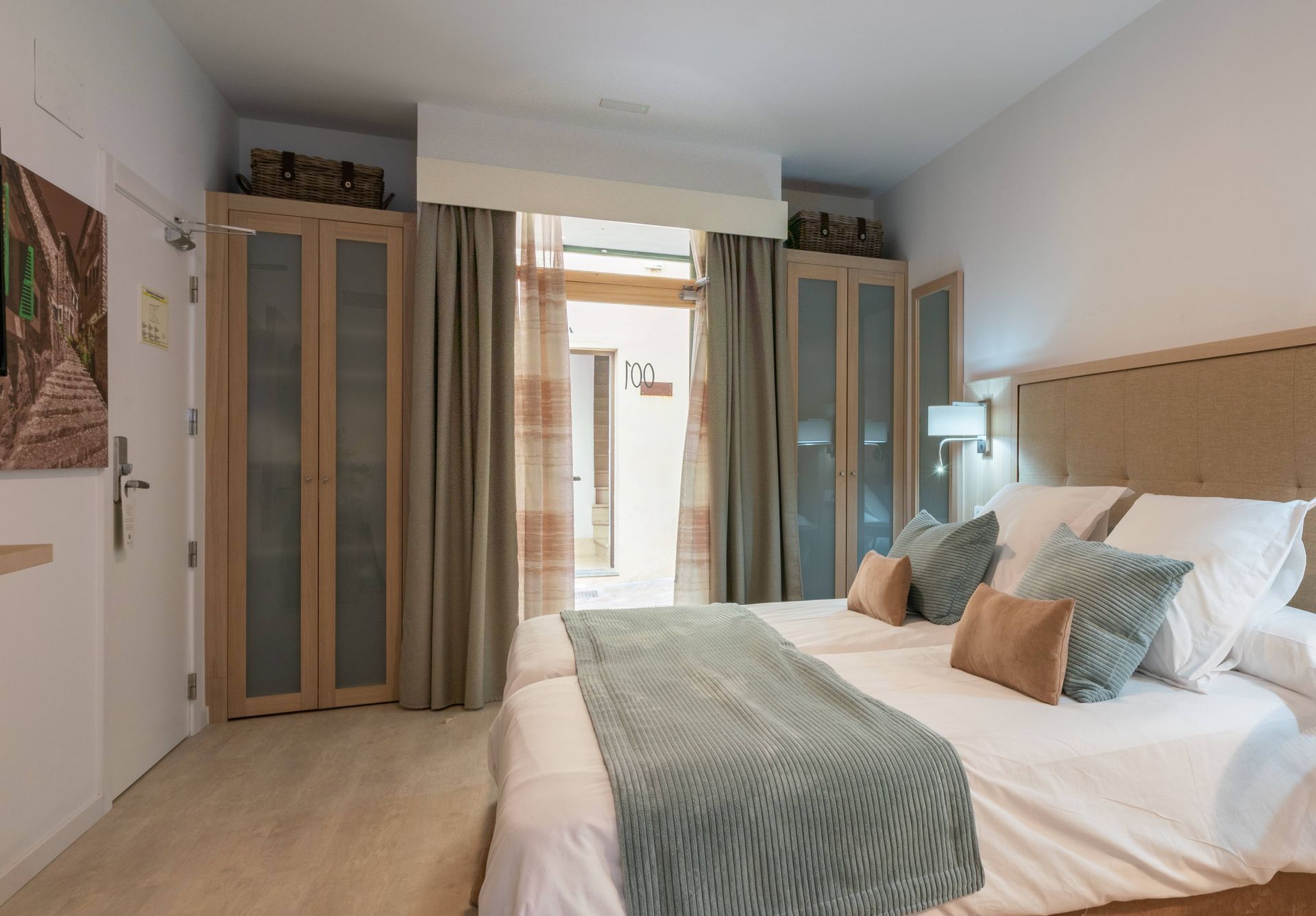 Bedroom with two beds, open doorway to outside, light-colored furniture, beige walls, and a blue and tan bedspread.