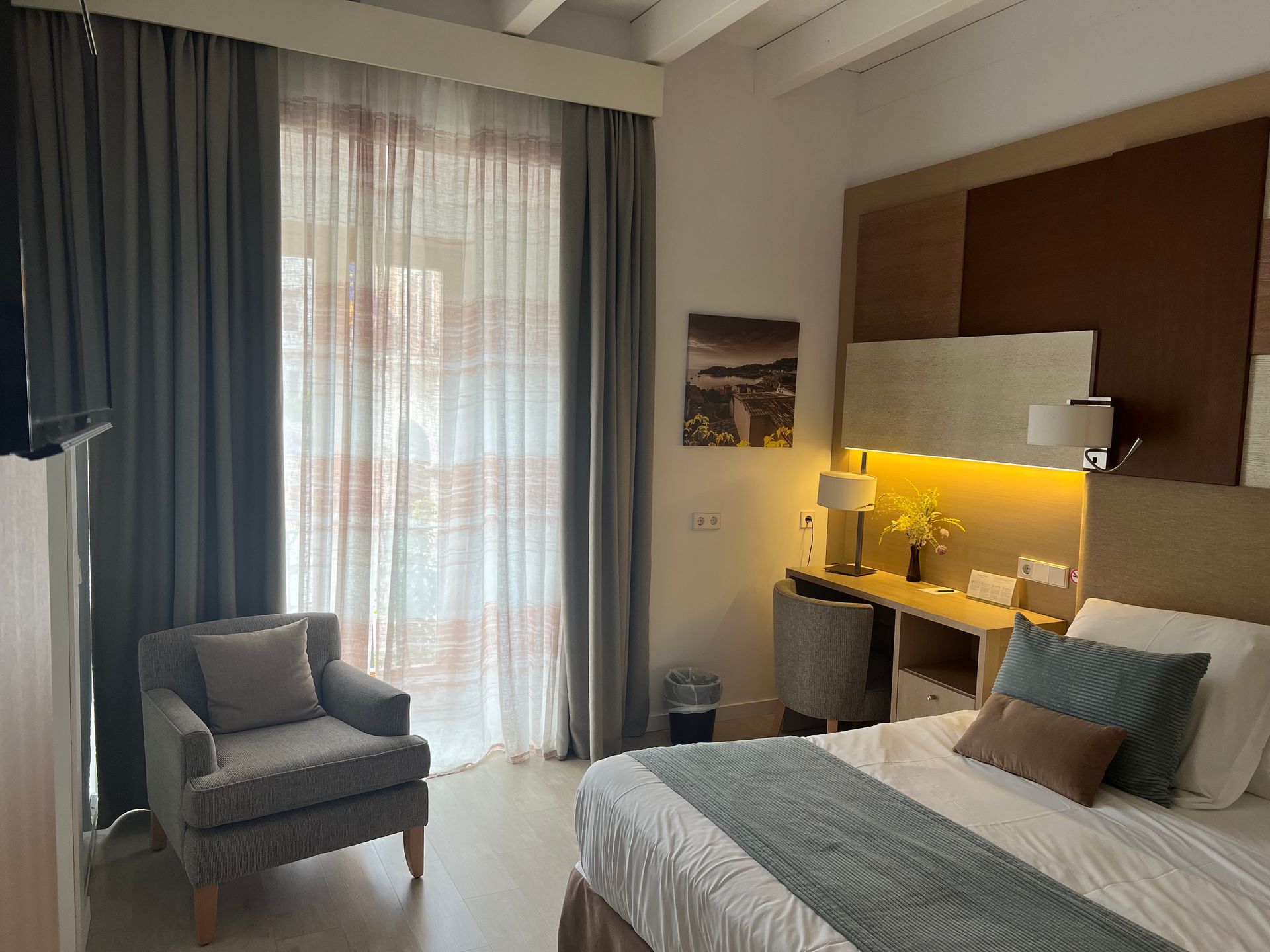Hotel room with bed, armchair, desk, and window with sheer curtains. Neutral colors, well-lit, cozy atmosphere.