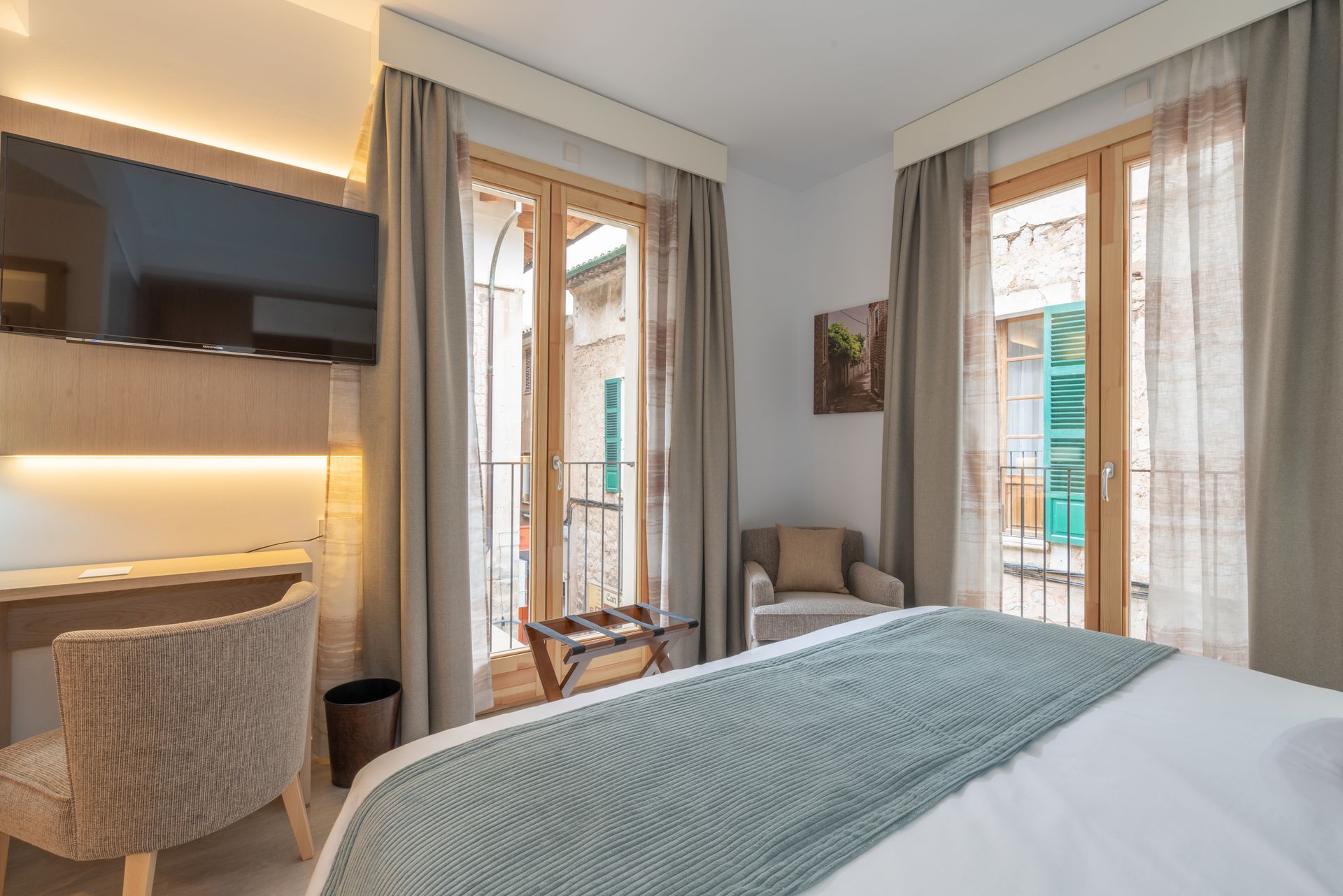 Hotel room with bed, desk, TV, and balcony overlooking an alleyway with colorful buildings.