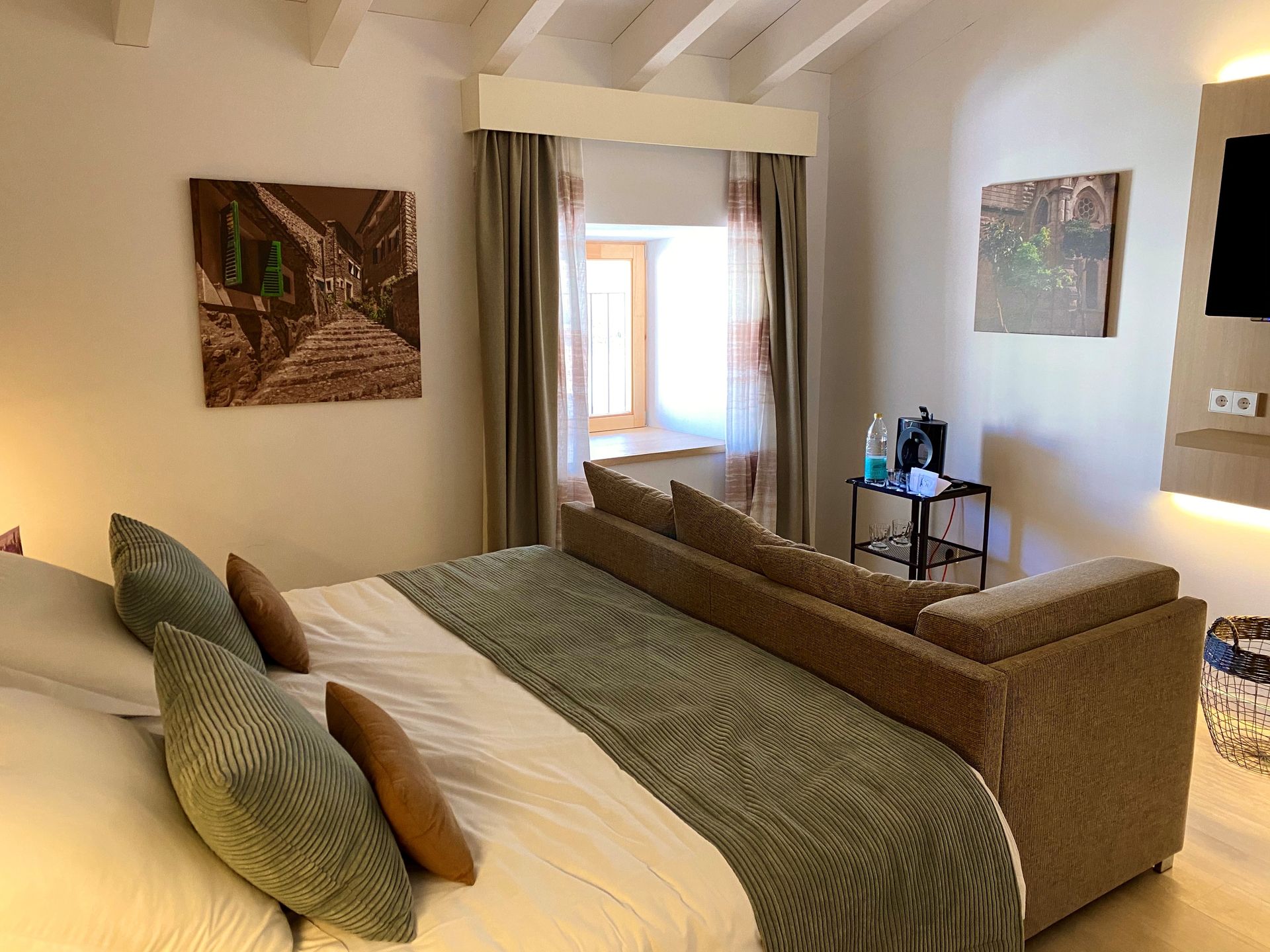 Bedroom with bed, sofa, artwork, window, and mini-bar. Neutral colors and wood beams.