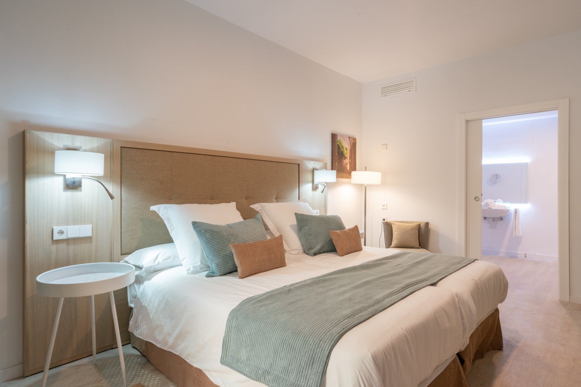 Bedroom with king-sized bed, neutral colors, accent pillows, side table, and doorway to a bathroom.