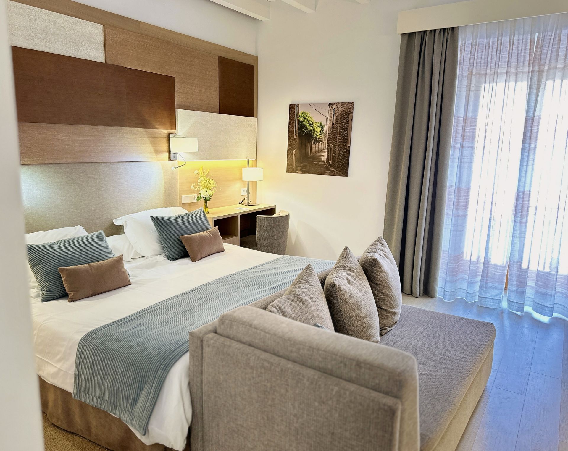 Bedroom with a large bed, chaise lounge, and window with sheer curtains. Neutral tones with brown and blue accents.