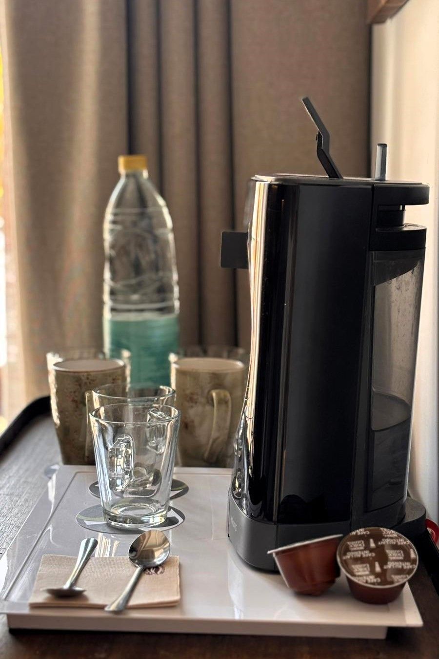 Coffee station: coffee maker Dolce Gusto, water bottle, glasses, spoon, fork, and coffee pods on a white tray.