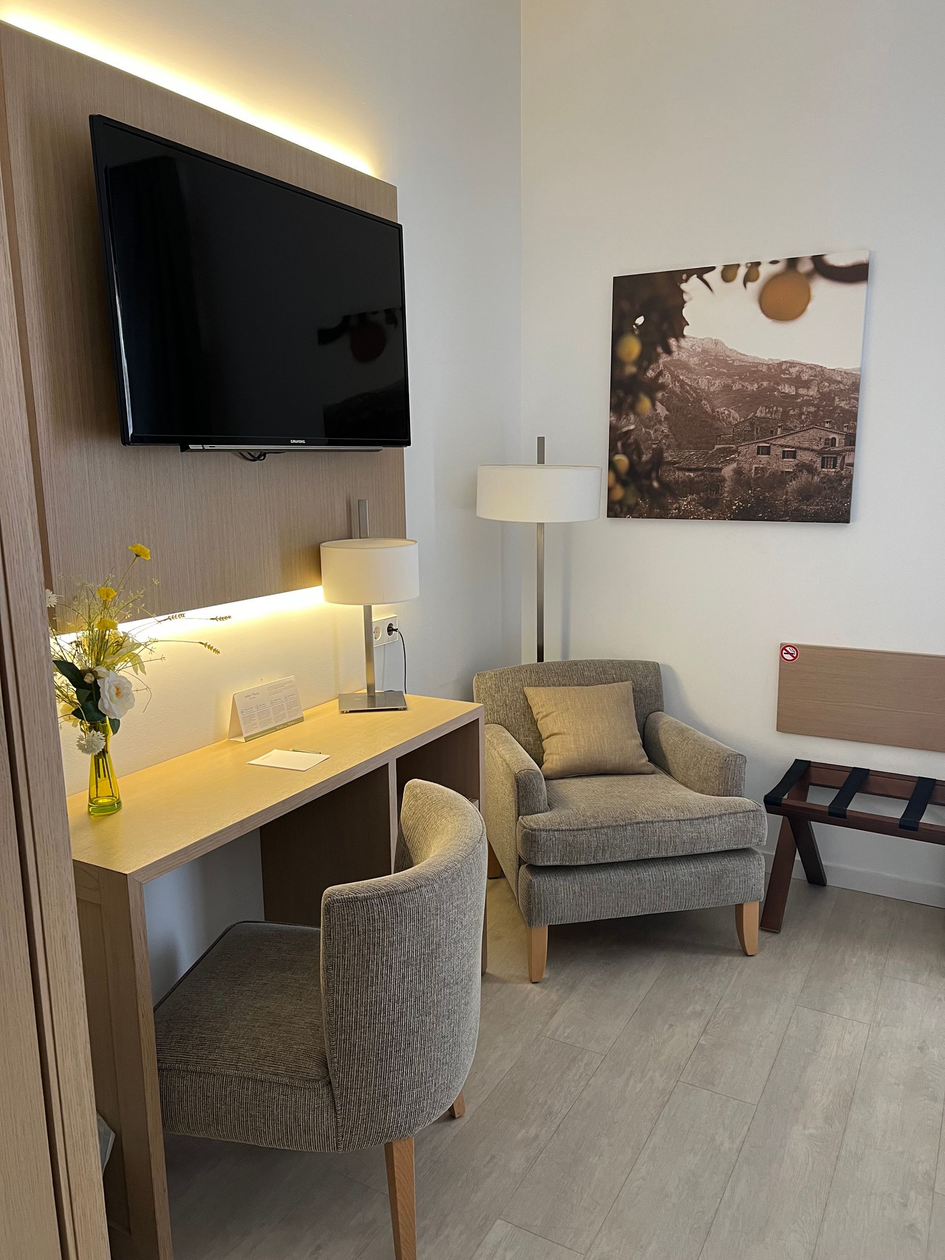 Cozy hotel room corner with a desk, armchair, TV, and artwork. Neutral tones, natural light.