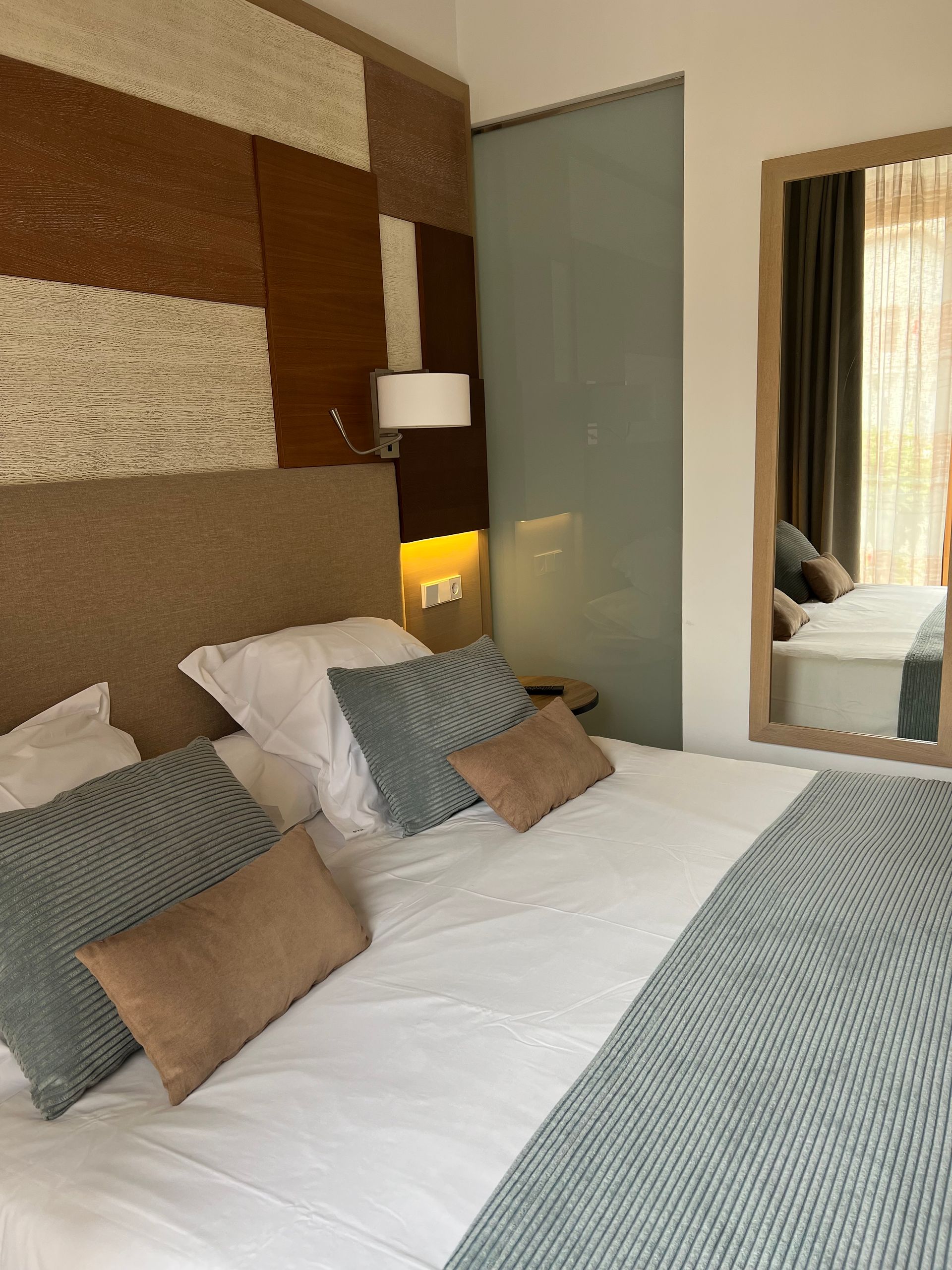 Bedroom with bed, decorative wall panels, mirror. Bed has pillows, a blue blanket. Beige, white and blue colors.