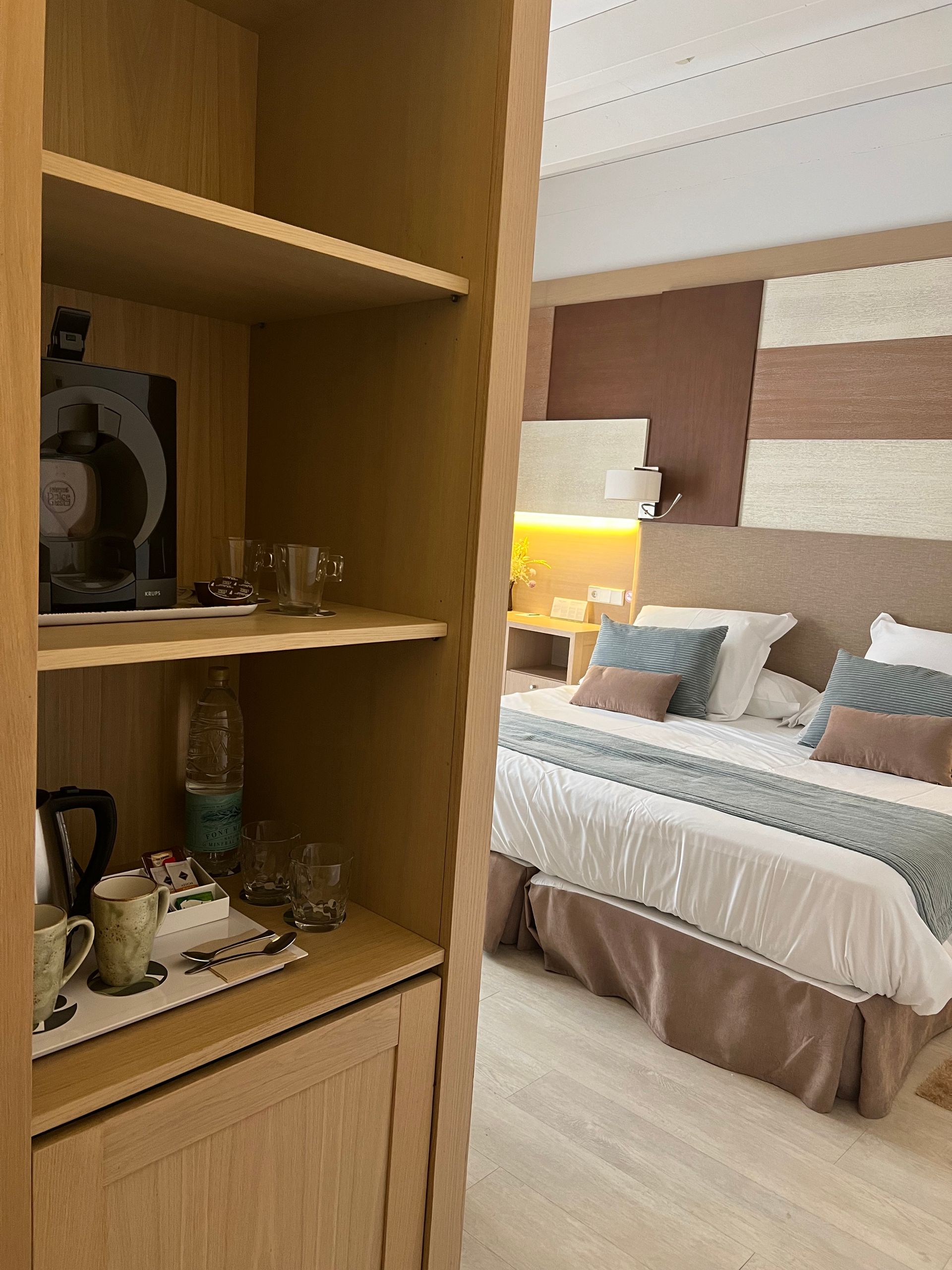 Wooden cabinet with coffee maker, glasses, and kettle. Bedroom with bed, pillows, and textured wall in the background.