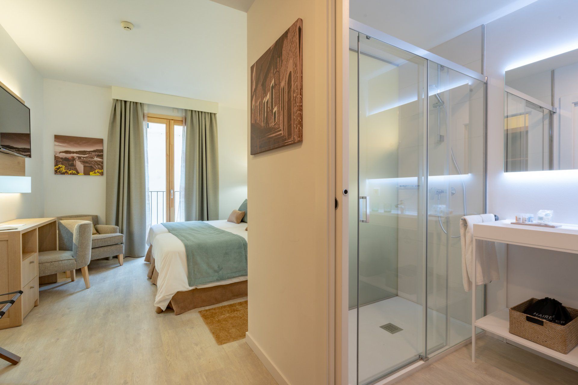 Hotel room with a view of the bed and a glass shower. Beige walls, wood floors, and a desk.
