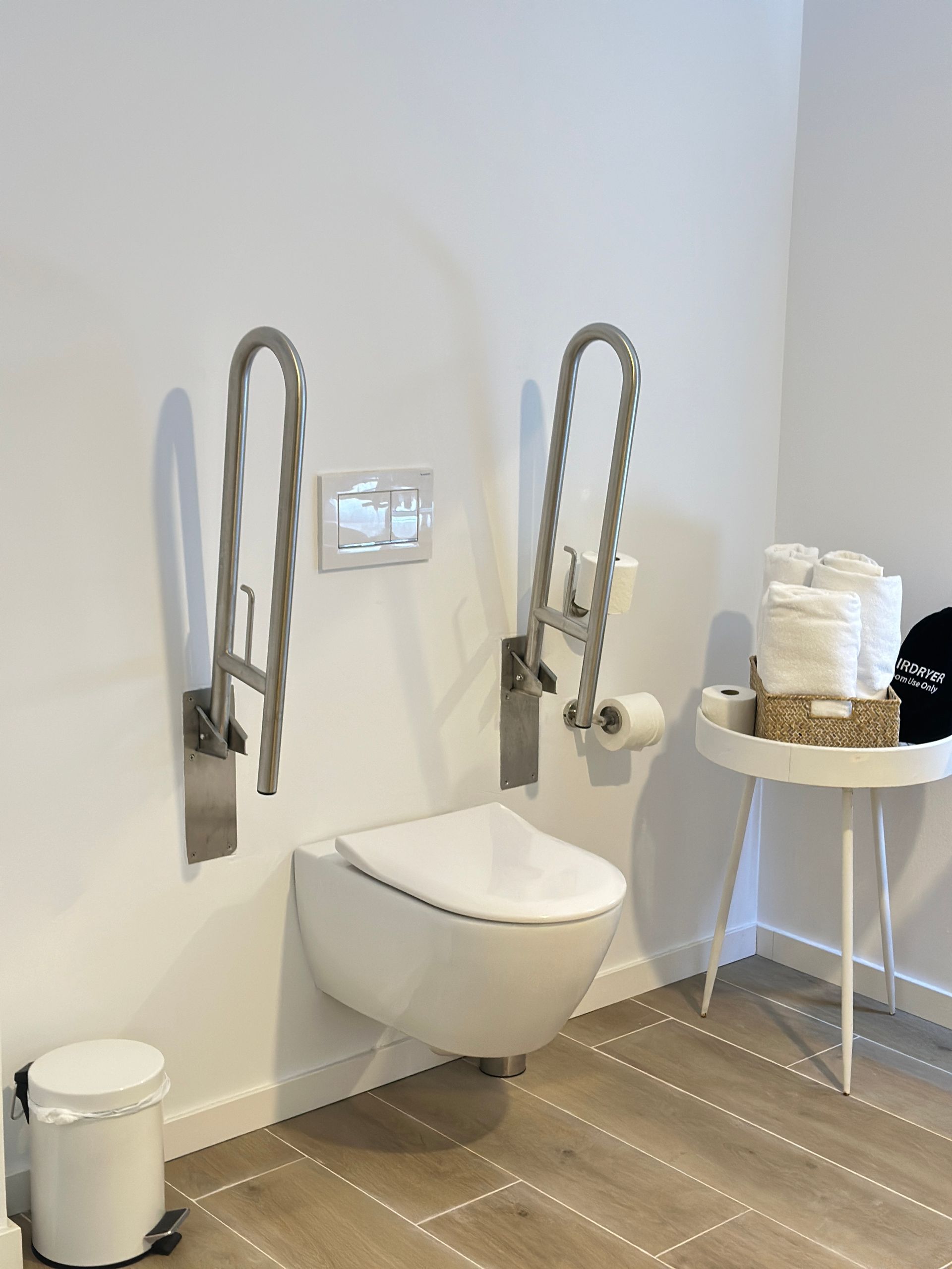 Accessible bathroom with wall-mounted toilet, grab bars, and a small table with towels.
