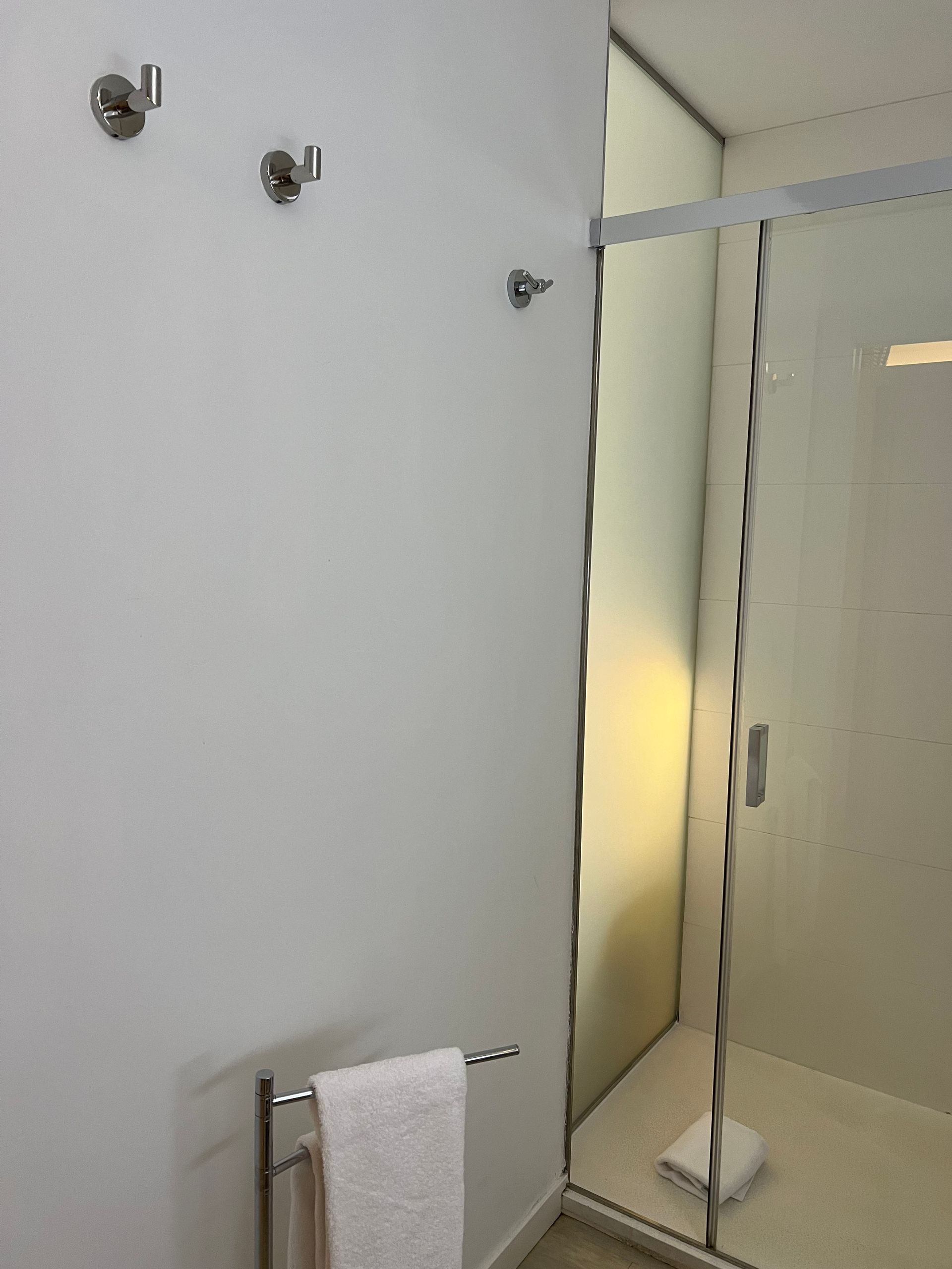 White bathroom with glass shower, towel rack, and three silver hooks on the wall.