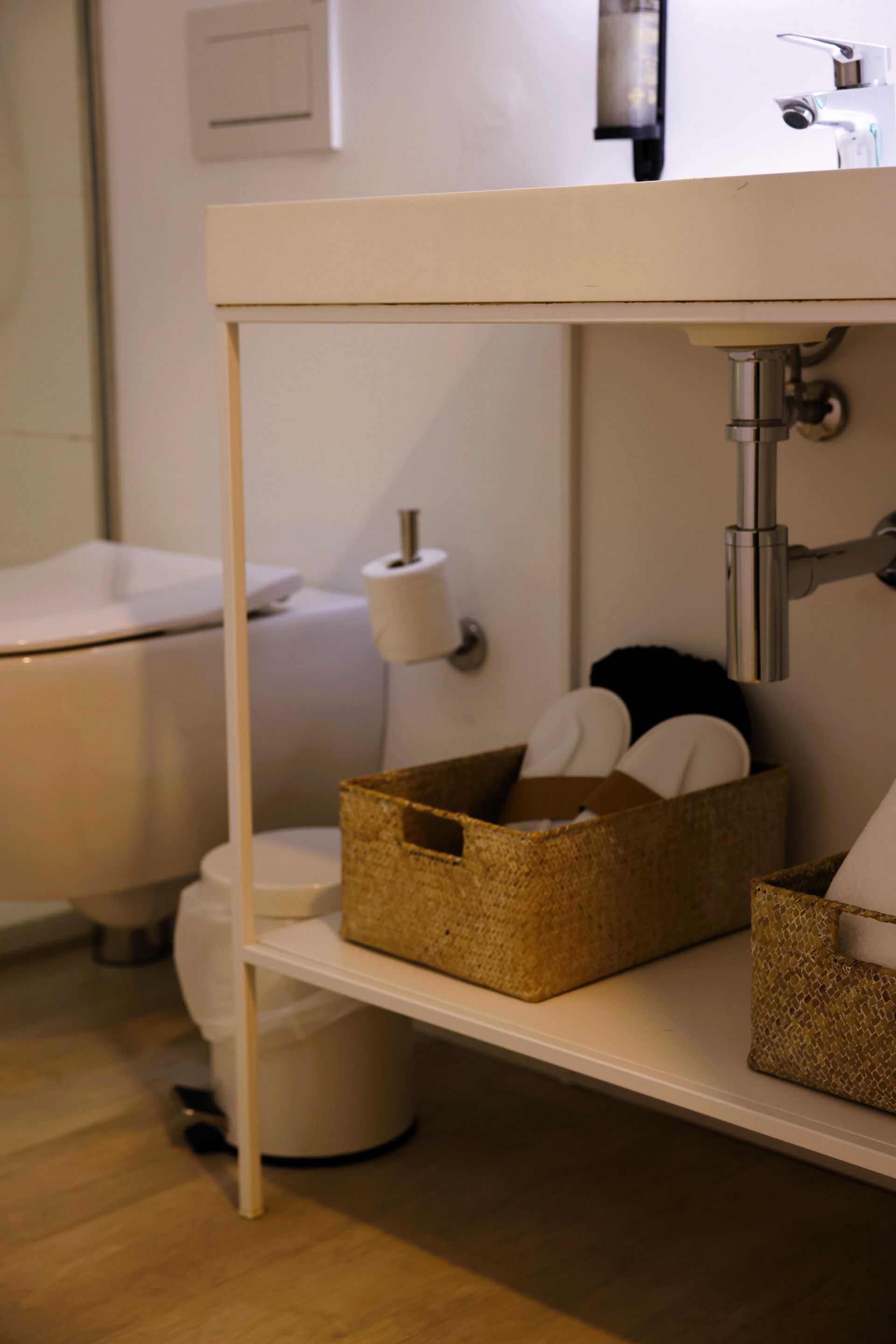 Bathroom with a white sink, toilet, and shelf containing slippers. Wicker baskets are used for storage.