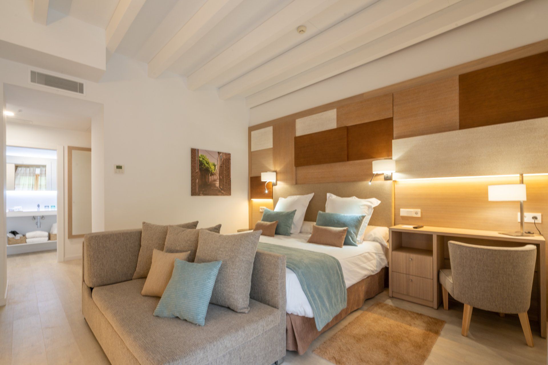 Hotel room with a bed, sofa, desk, and bathroom entrance. Neutral tones, cozy atmosphere.