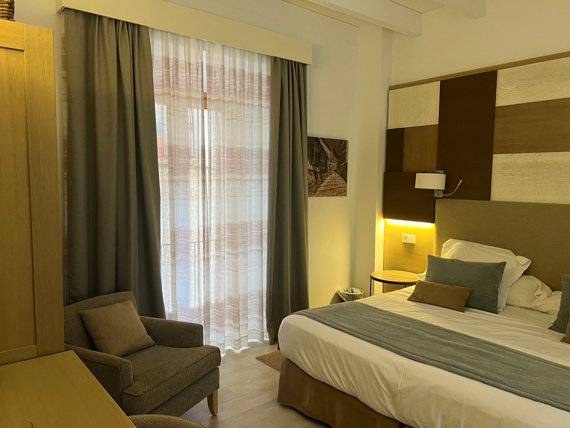 Bedroom with a bed, armchair, and window with curtains. Neutral colors and warm lighting.