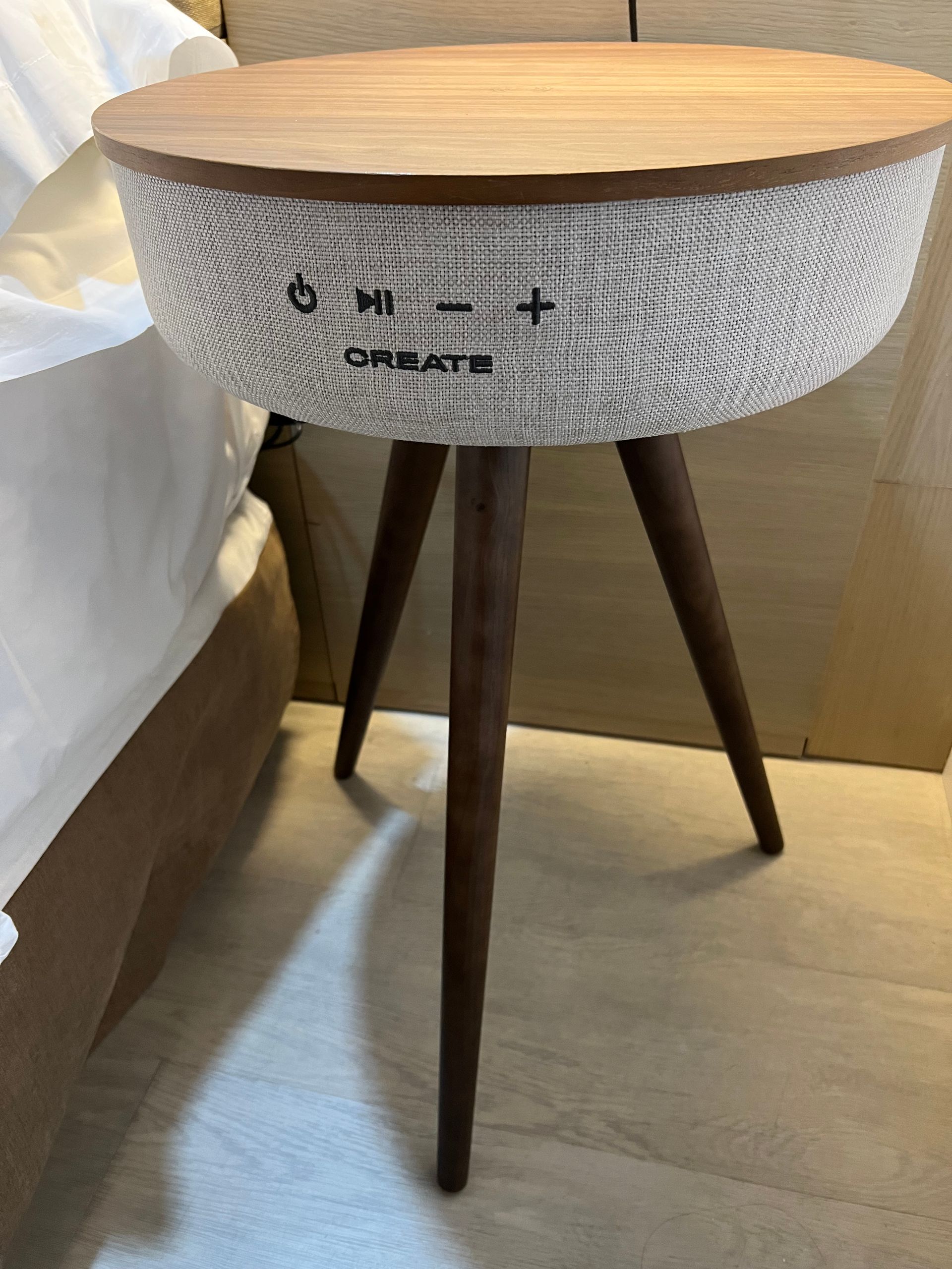 A round, wooden-topped side table with a fabric speaker, three legs, and controls. Create